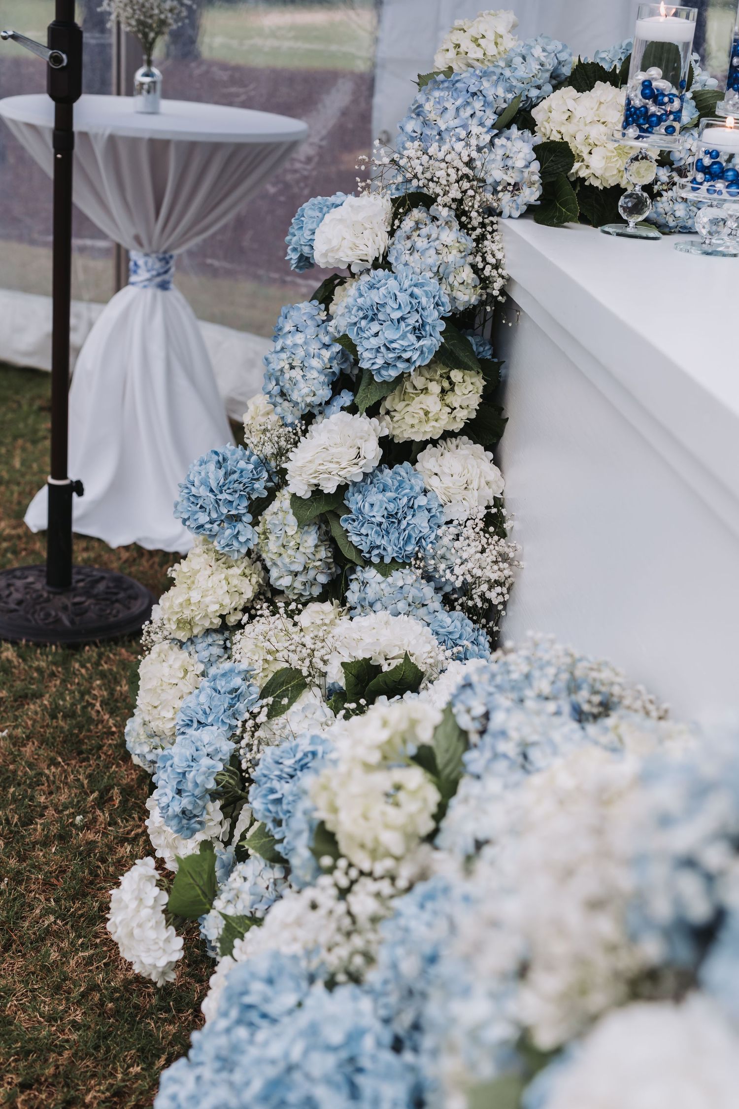 Cascading blue and white hydrangea floral arrangement decorating the edge of a wedding reception table outdoors.
