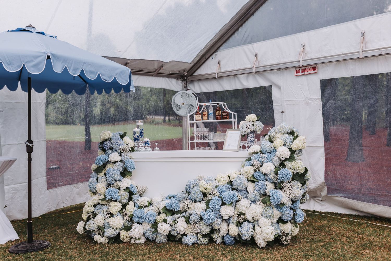Elegant blue and white hydrangea floral bar display under striped umbrella at outdoor wedding reception tent.