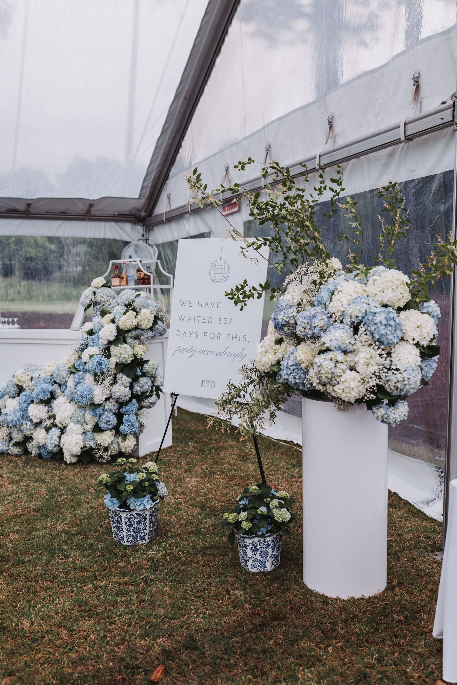 Blue and white hydrangea floral displays on white pedestals inside a clear wedding tent with welcome signage.