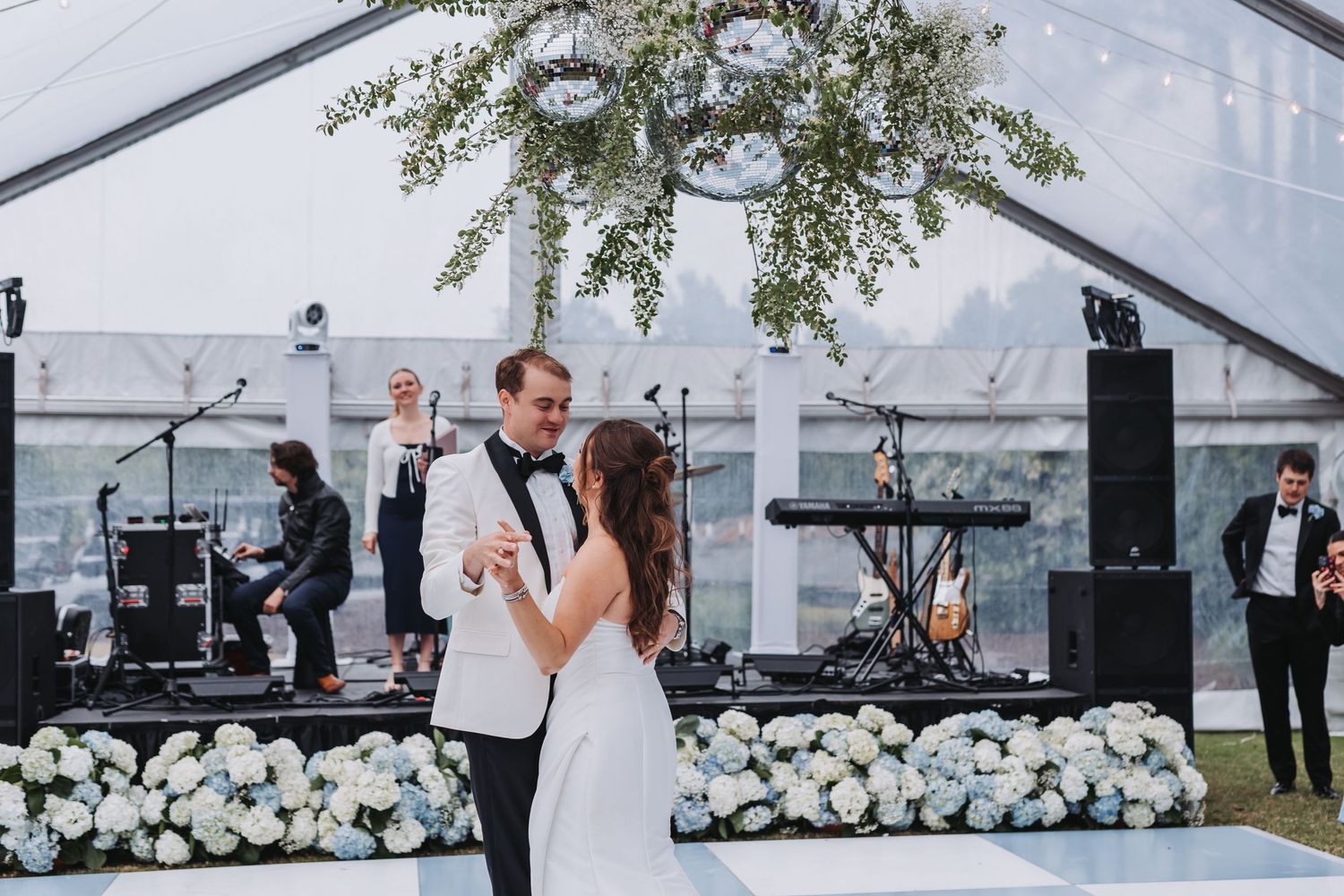 Bride and groom share first dance under floral chandelier in clear tent with live band performing.
