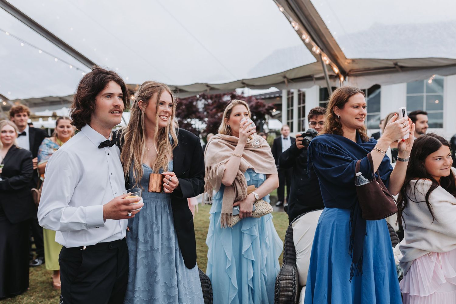 Wedding guests in formal attire watch ceremony outdoors under a clear tent on a cloudy day.