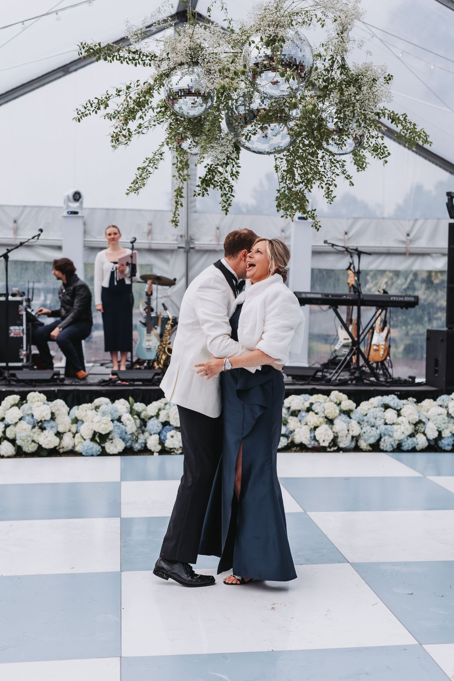 Couple shares first dance on checkered floor under floral chandelier at elegant wedding reception.