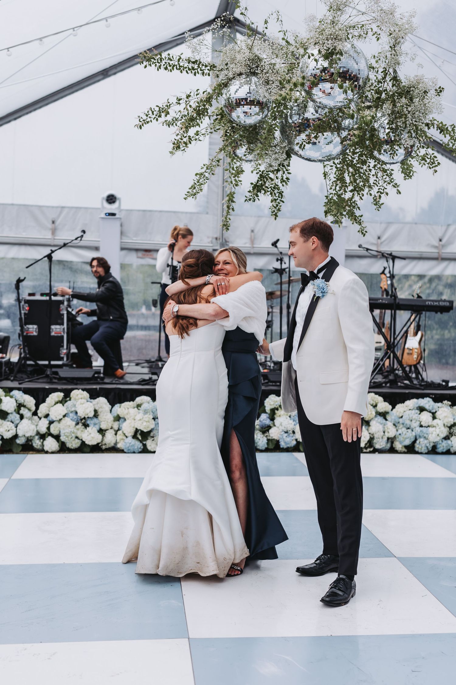 Bride hugs guest on checkered dance floor as groom watches, with hydrangea floral wall and live band backdrop.