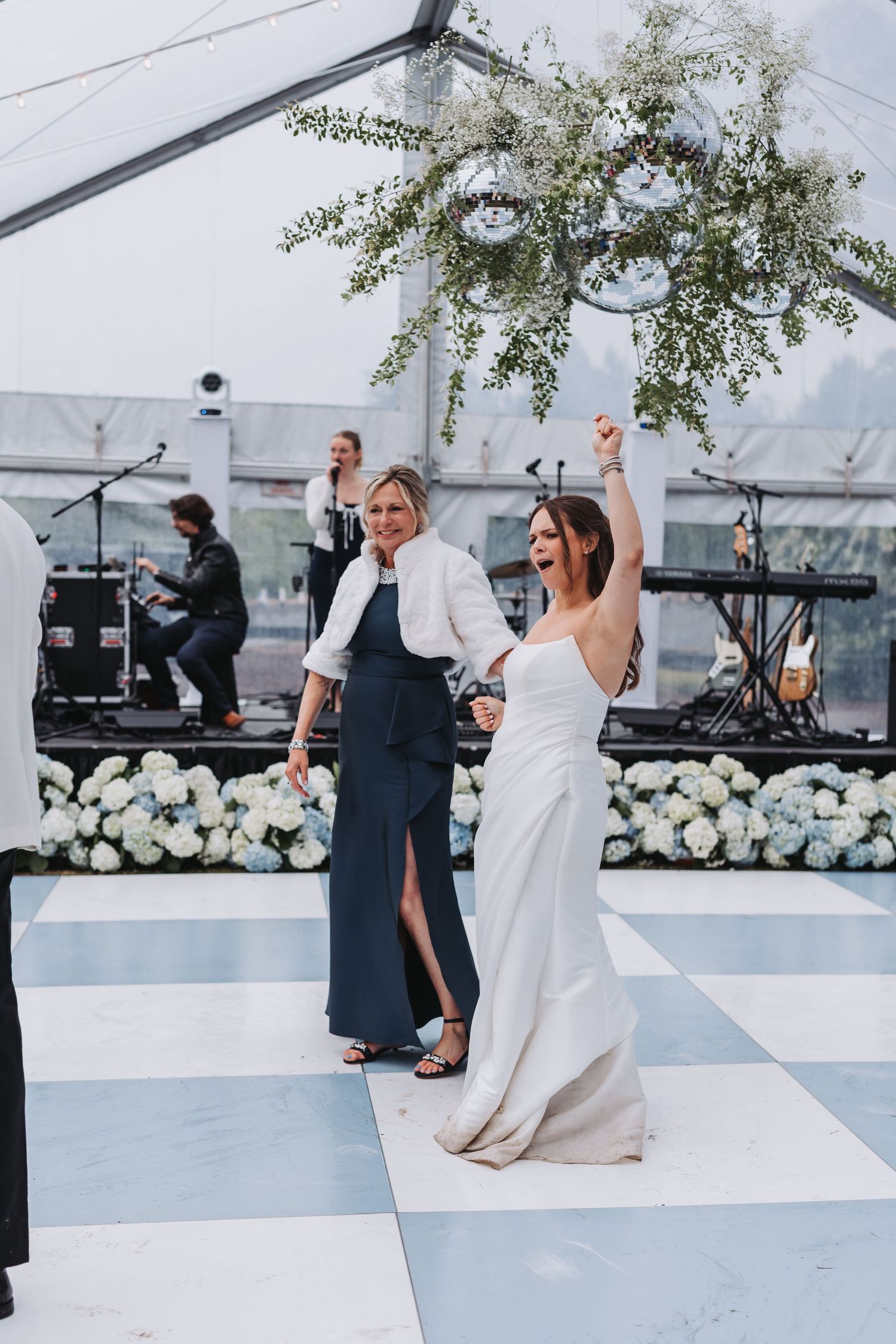 Bride in white strapless gown raises arm joyfully on checkered dance floor under floral chandelier at wedding reception.