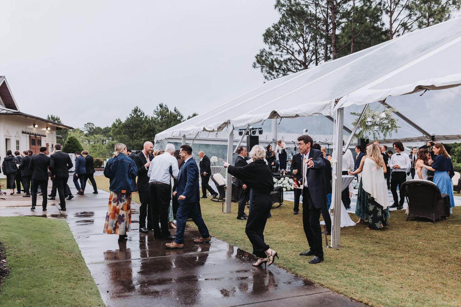 Wedding guests mingling on wet pavement outside a white tent during a rainy outdoor reception.