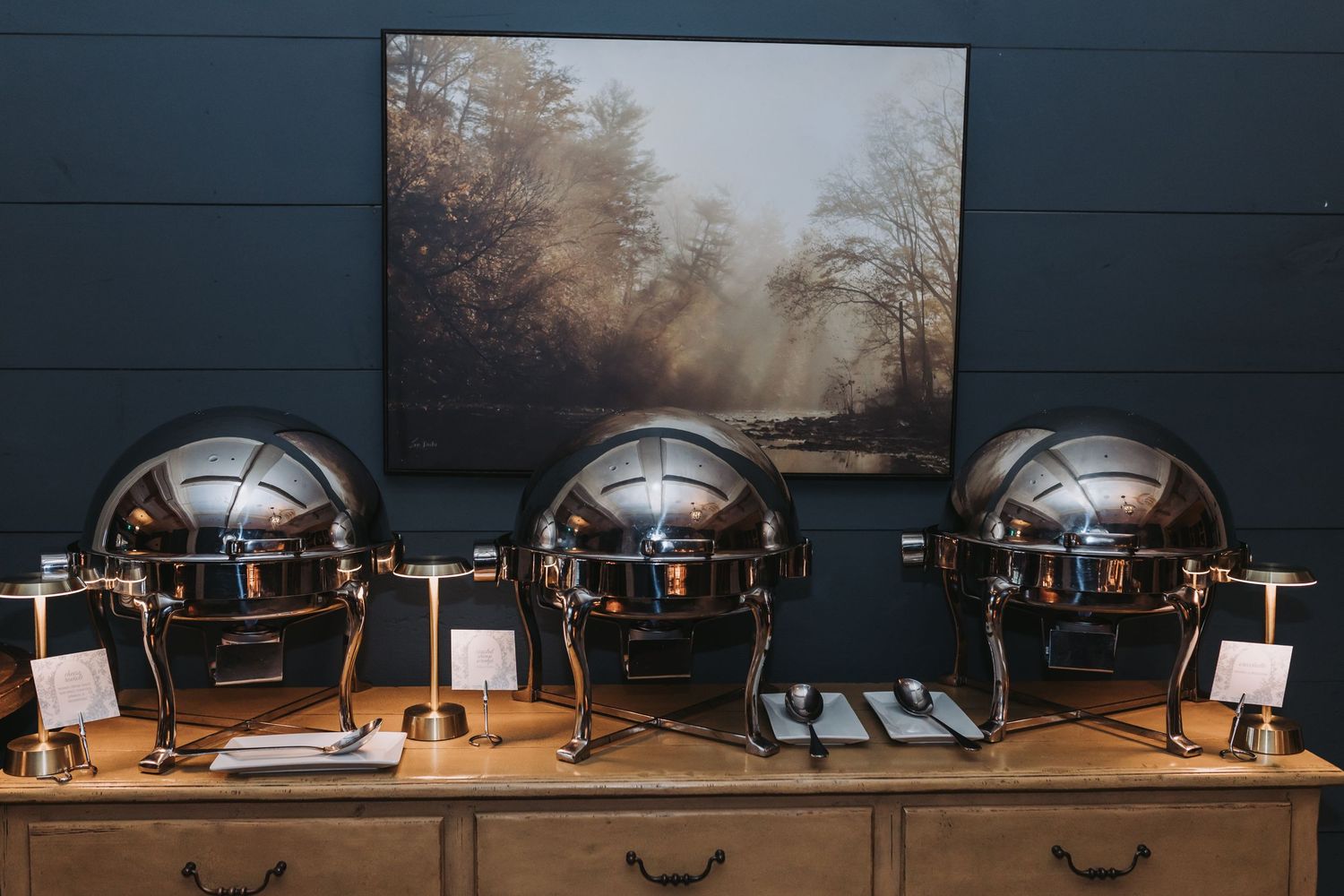Three polished silver dome chafing dishes arranged on a wooden buffet table at a catered wedding reception.