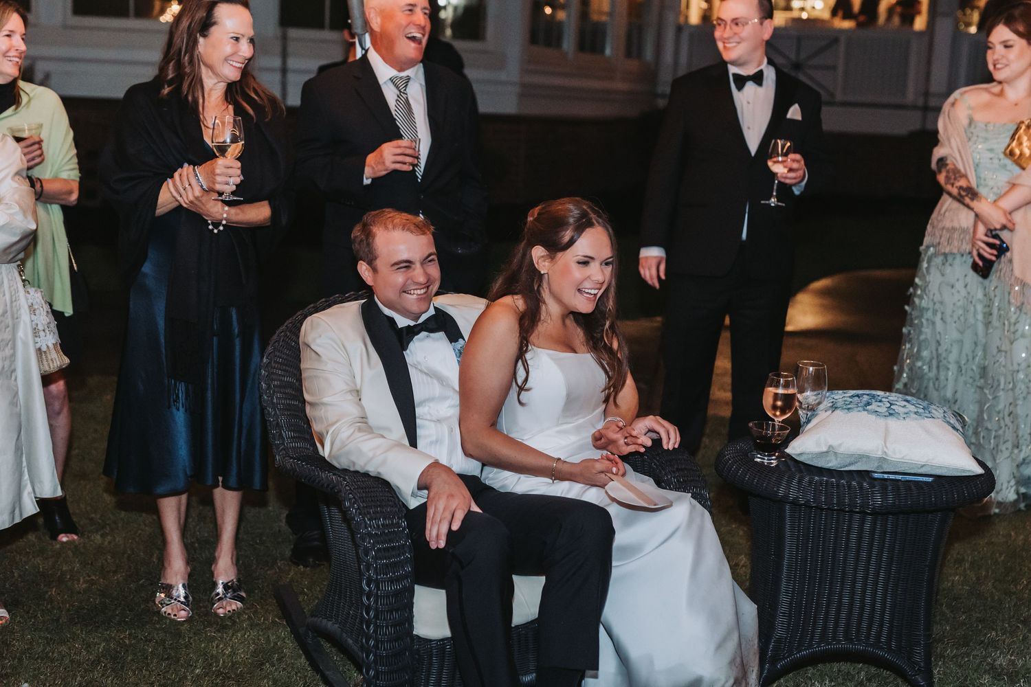 Bride and groom seated together smiling surrounded by elegantly dressed wedding guests outdoors at night.