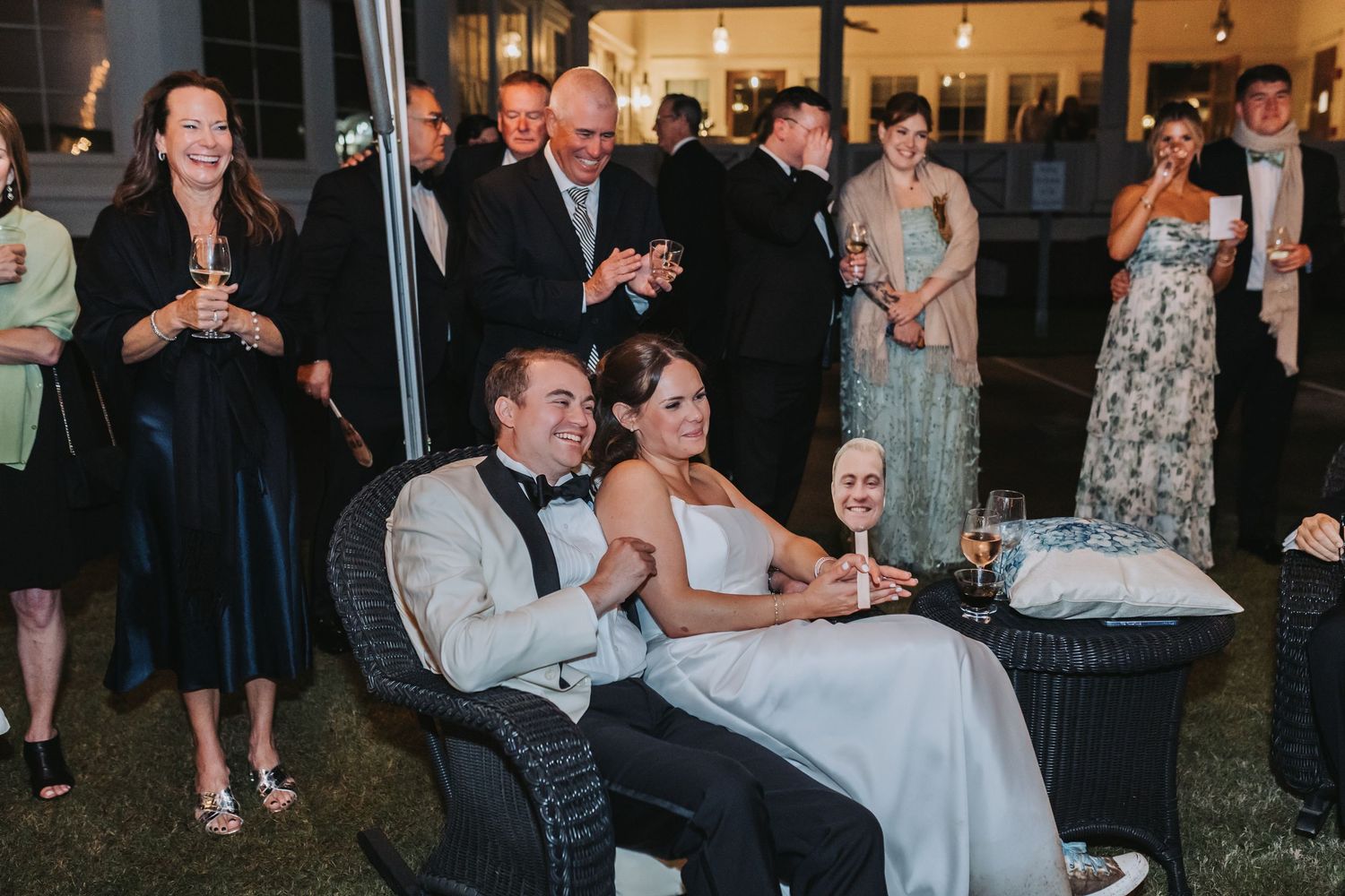 Bride and groom relax in wicker chairs surrounded by wedding guests socializing outdoors at evening reception.