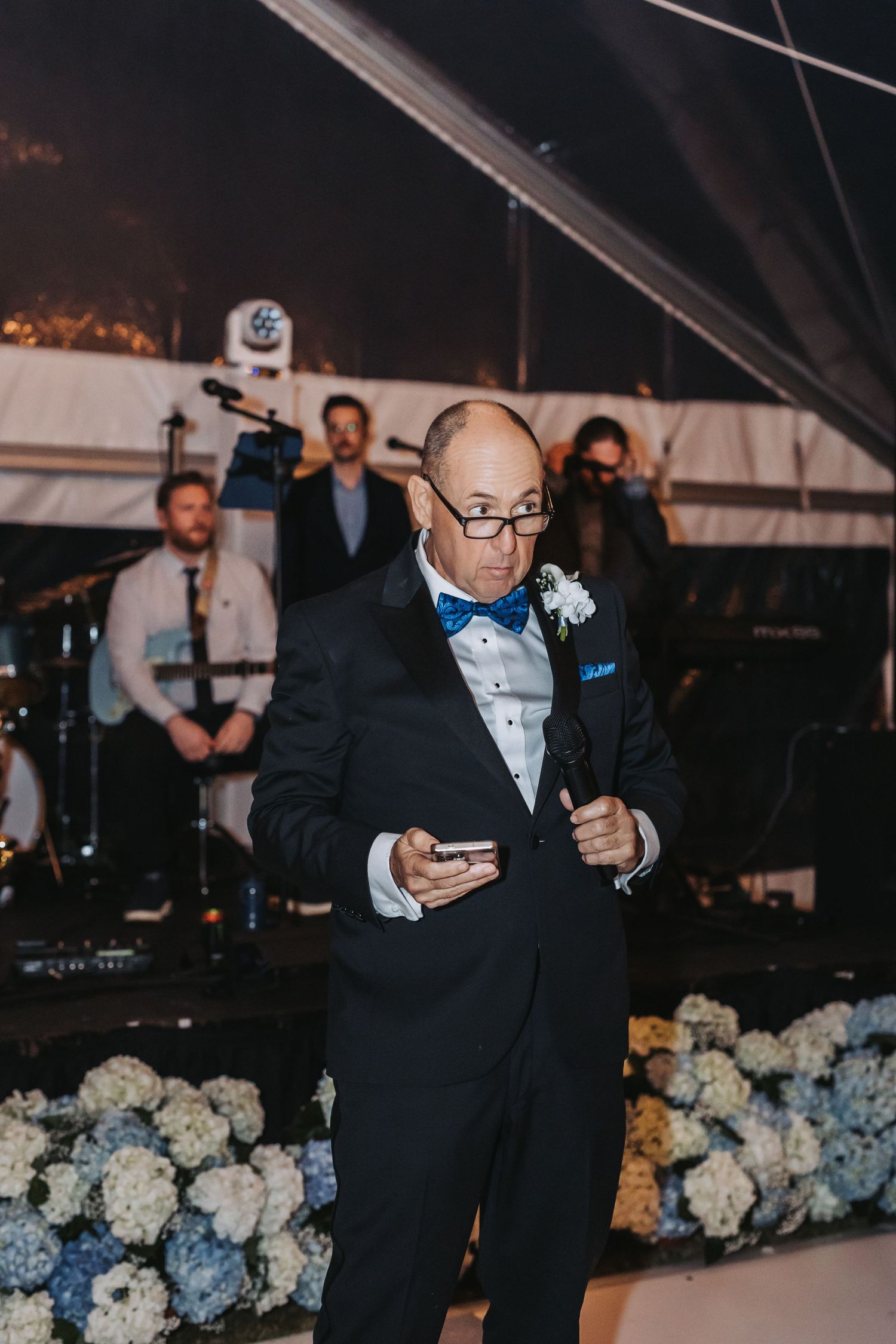 Dapper wedding guest in black tuxedo with blue bow tie delivers speech on dance floor at elegant reception.