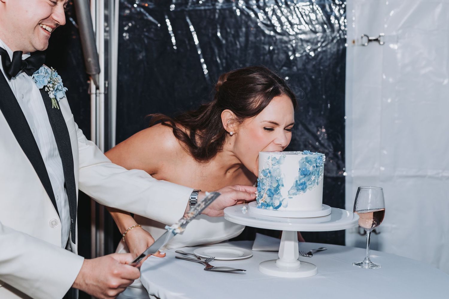 Bride playfully leans in to bite blue marble wedding cake as groom holds cake knife at reception.