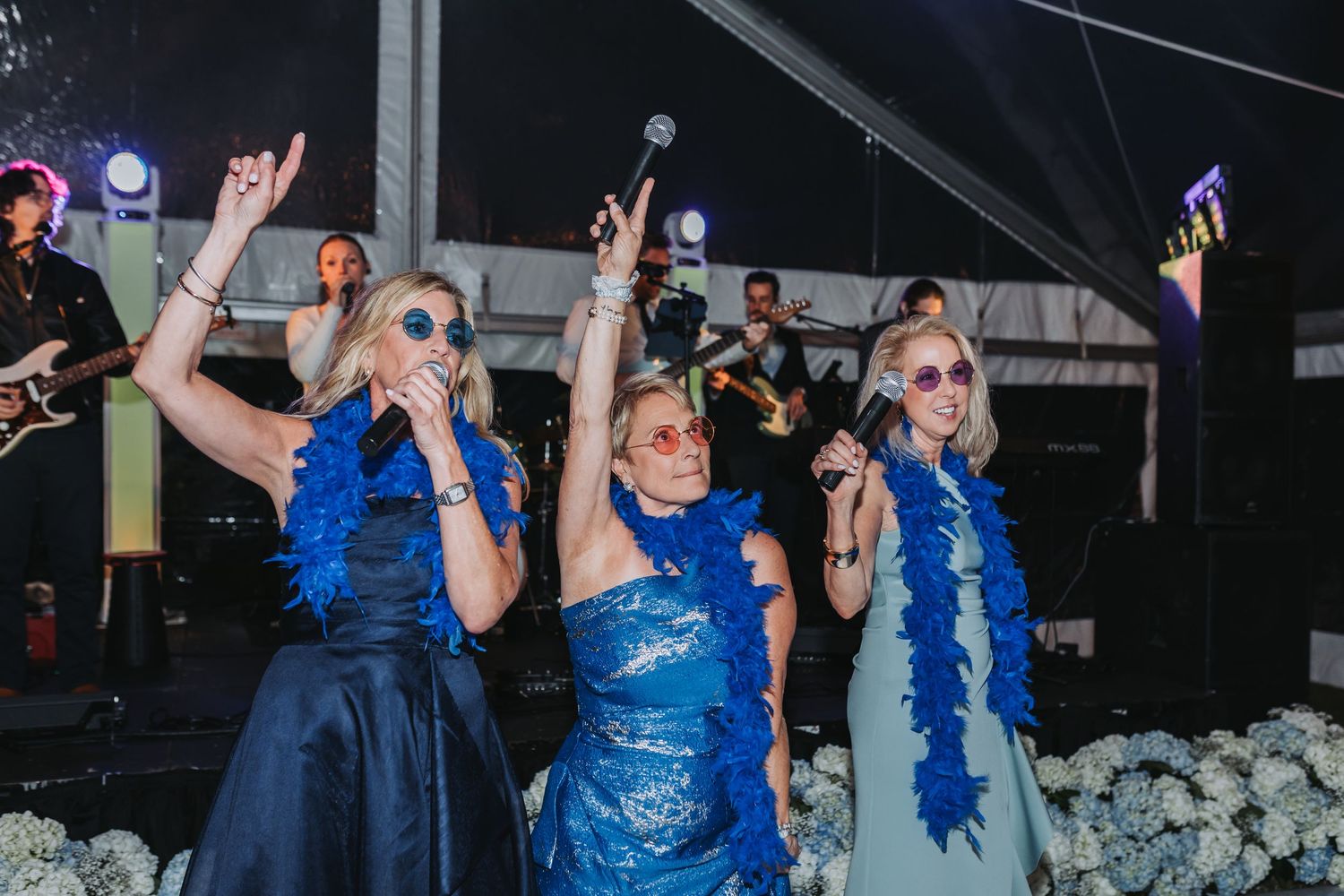 Three women in blue dresses with feather boas and sunglasses sing into microphones at a wedding reception.