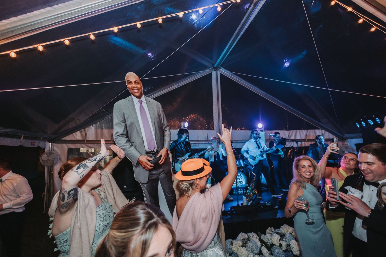 Tall man in gray suit stands on dance floor surrounded by cheering guests at lively wedding reception.