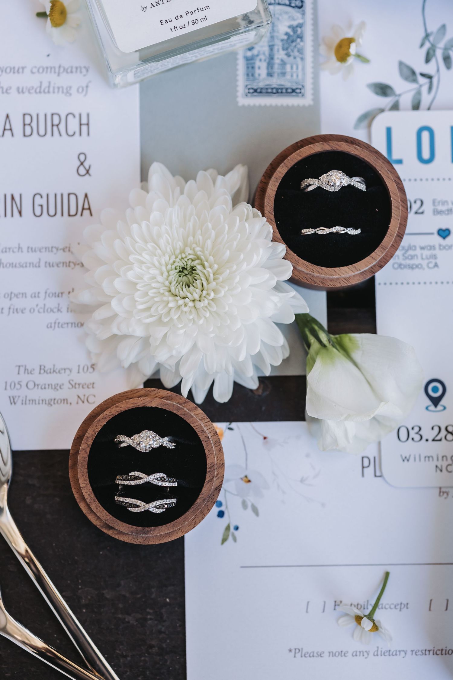 Wedding rings displayed in wooden velvet boxes beside white flowers and elegant invitation suite on dark surface.