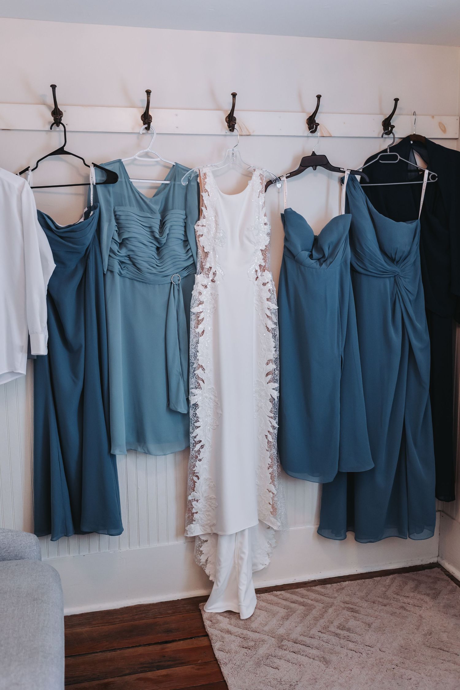 Blue bridesmaid dresses and white lace wedding gown hanging on wall hooks in a bridal suite.