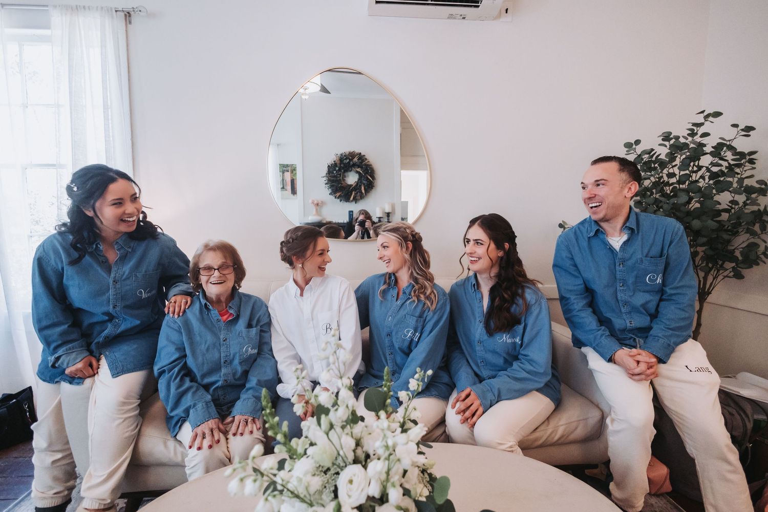 A joyful wedding party in matching denim shirts sits together on a couch, laughing and smiling around a floral arrangement.