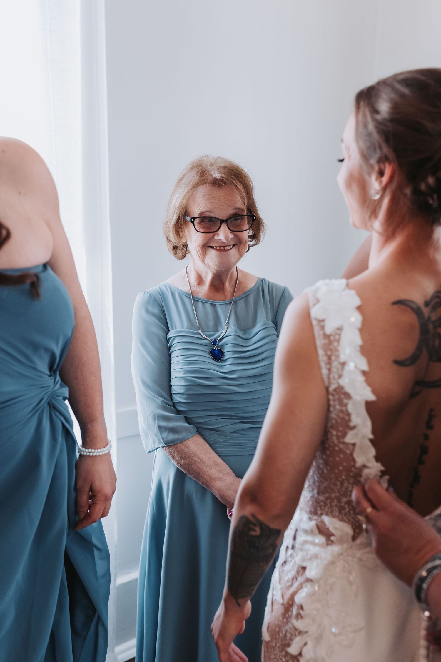 Smiling older woman in blue dress admiring bride in lace wedding gown.