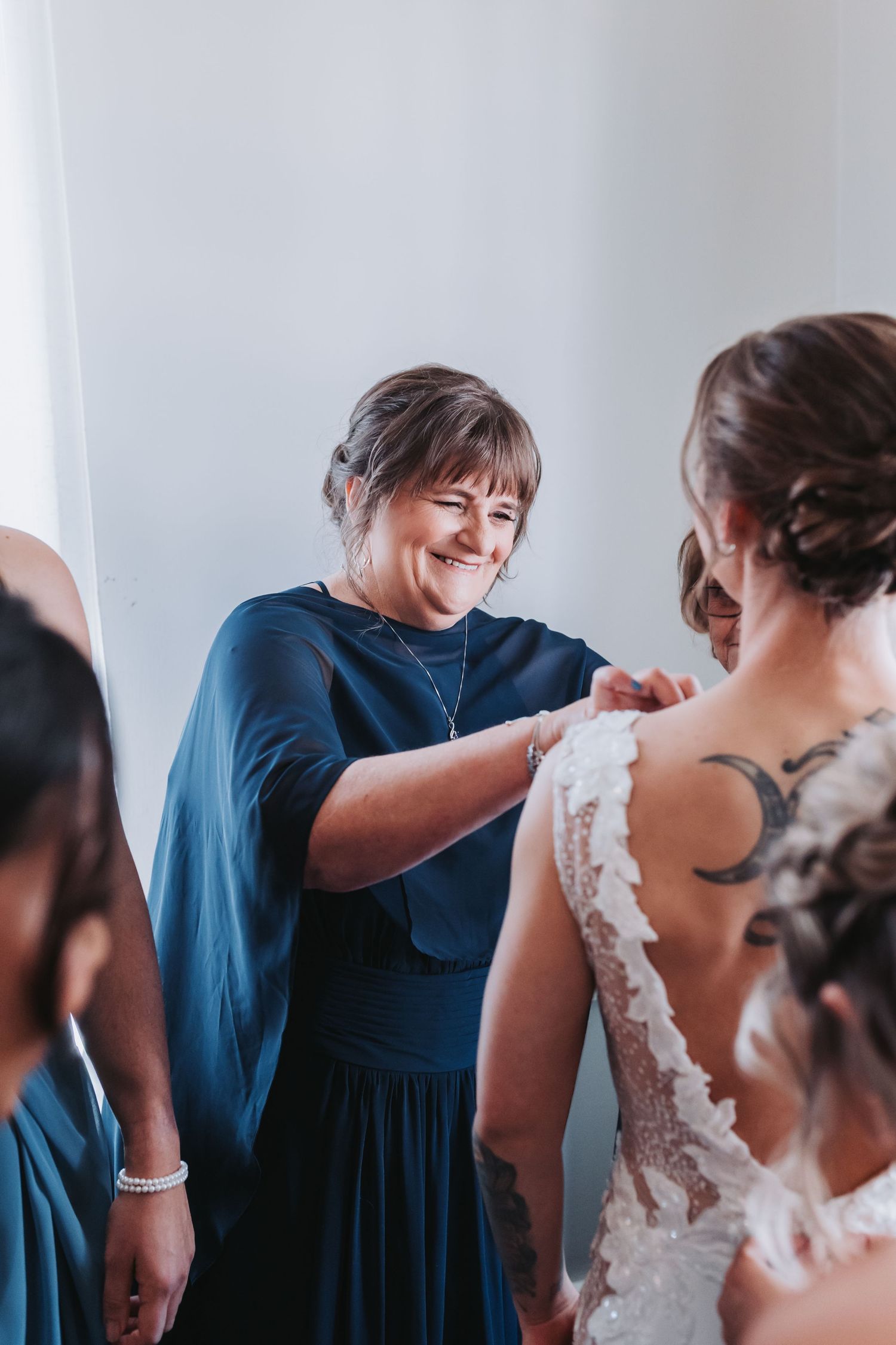 Smiling woman in navy dress buttoning the back of a brides lace wedding gown.