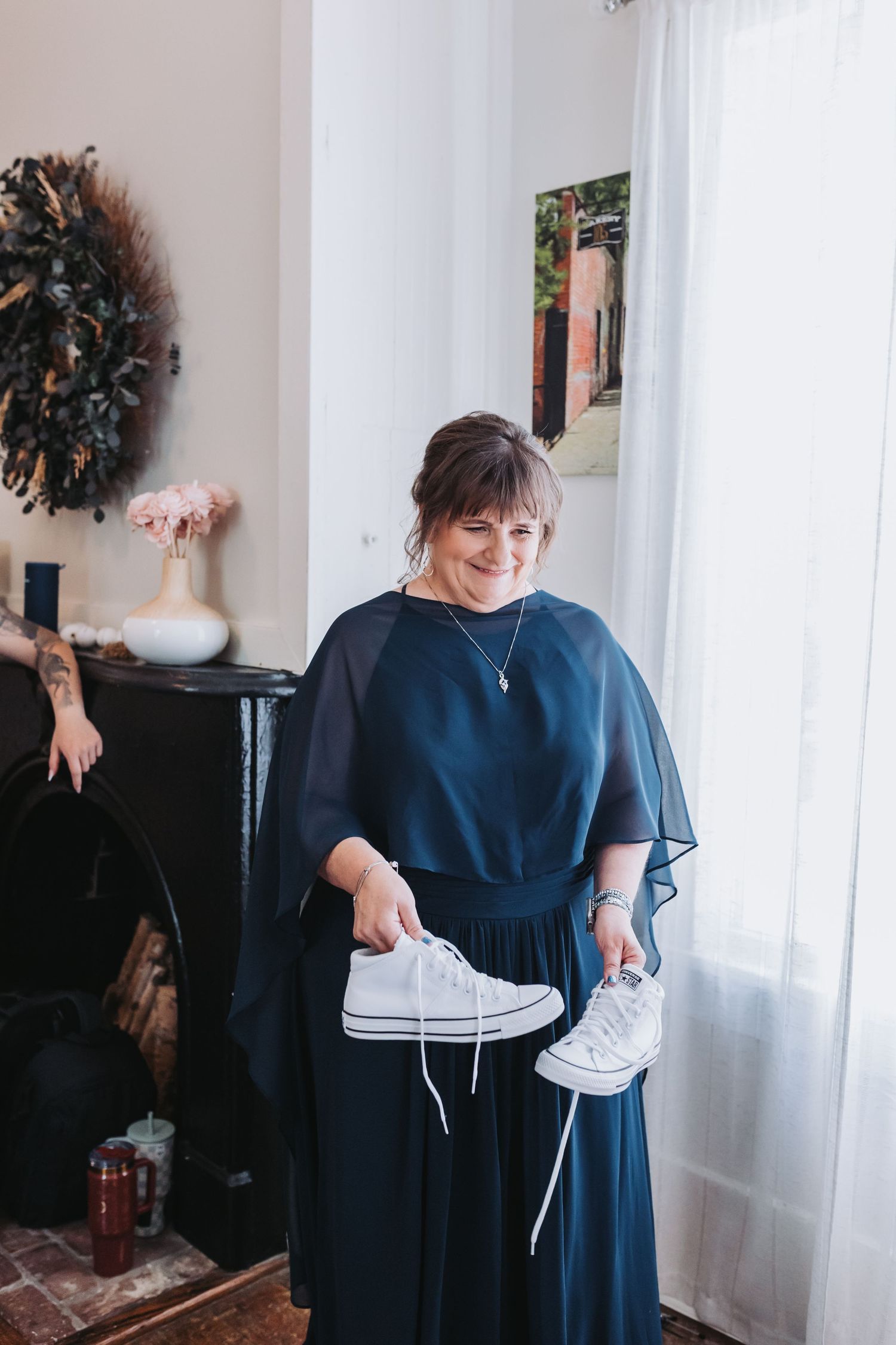 Woman in navy blue dress holding white Converse sneakers, smiling near a bright window in an elegant room.