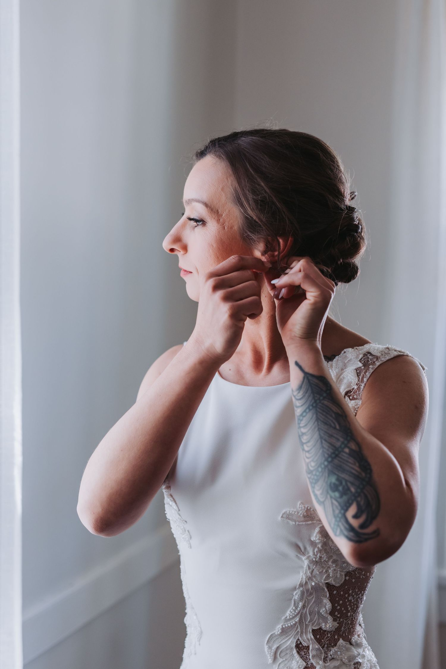 Tattooed bride in white lace wedding dress putting on earrings by a bright window.