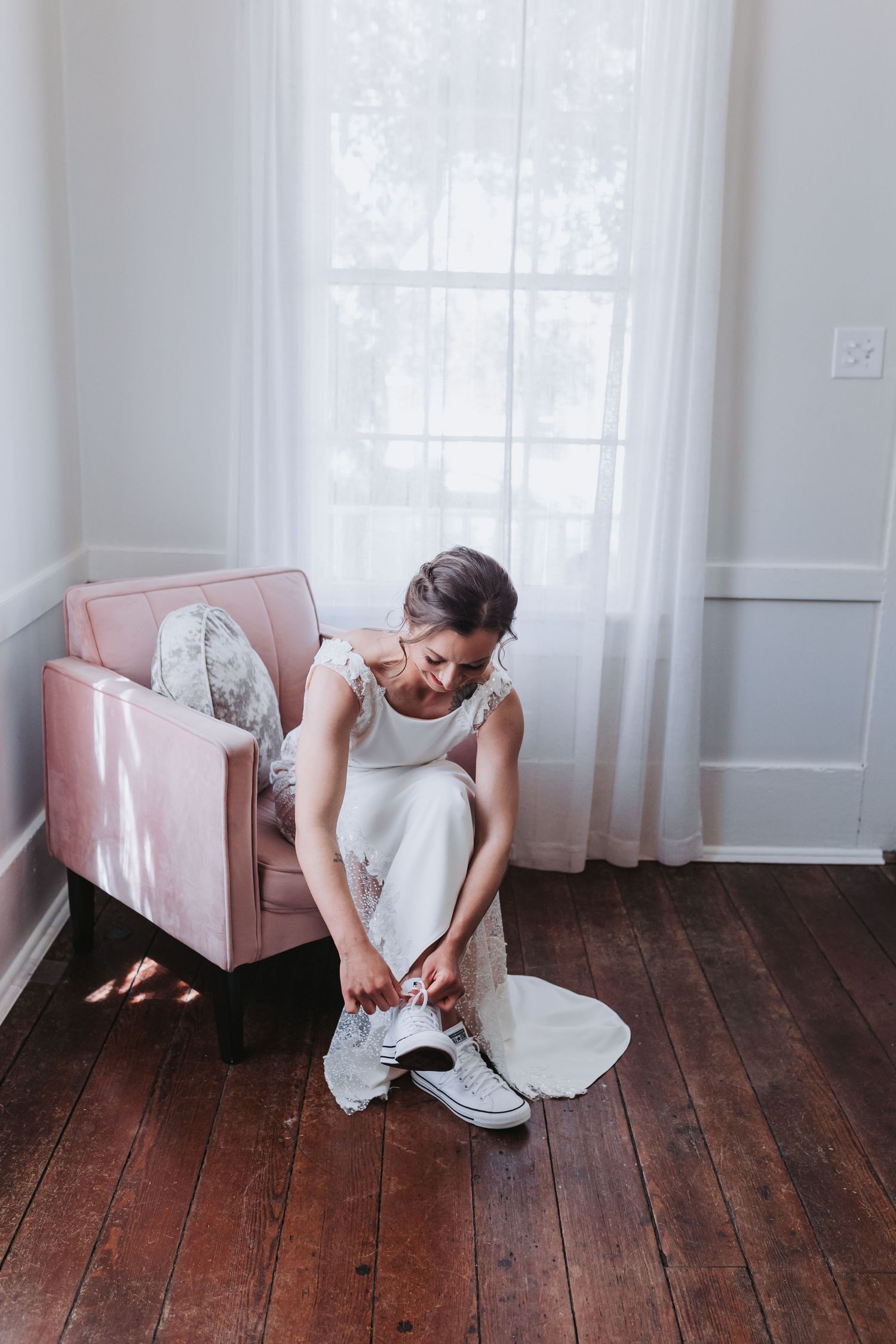 Bride in a white wedding gown tying her Converse sneakers while seated by a bright window.