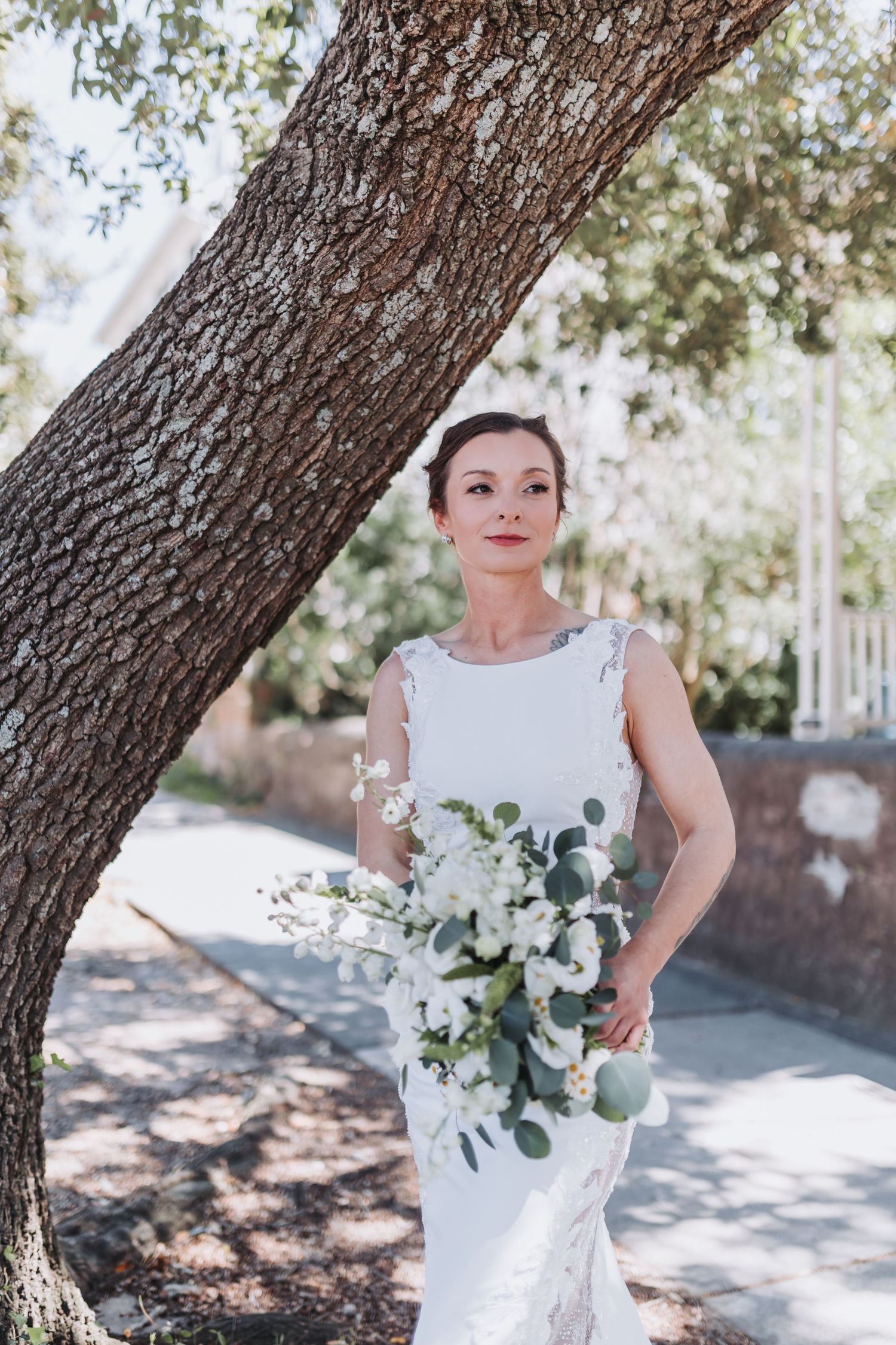 Elegant bride in sleeveless lace gown holds white eucalyptus bouquet while posing beside large tree trunk.