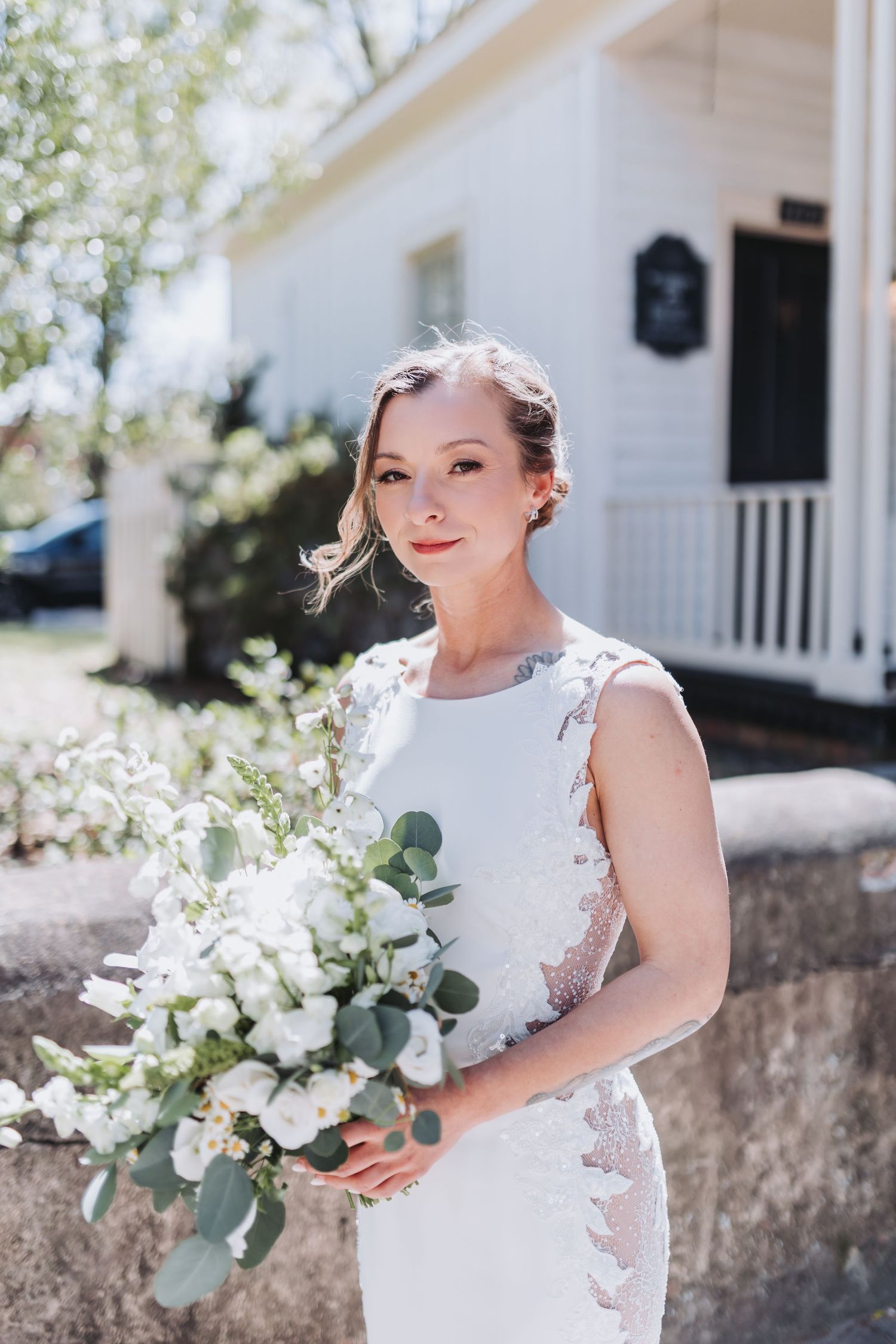 Elegant bride in white lace gown holding white and green bouquet outdoors on sunny wedding day.