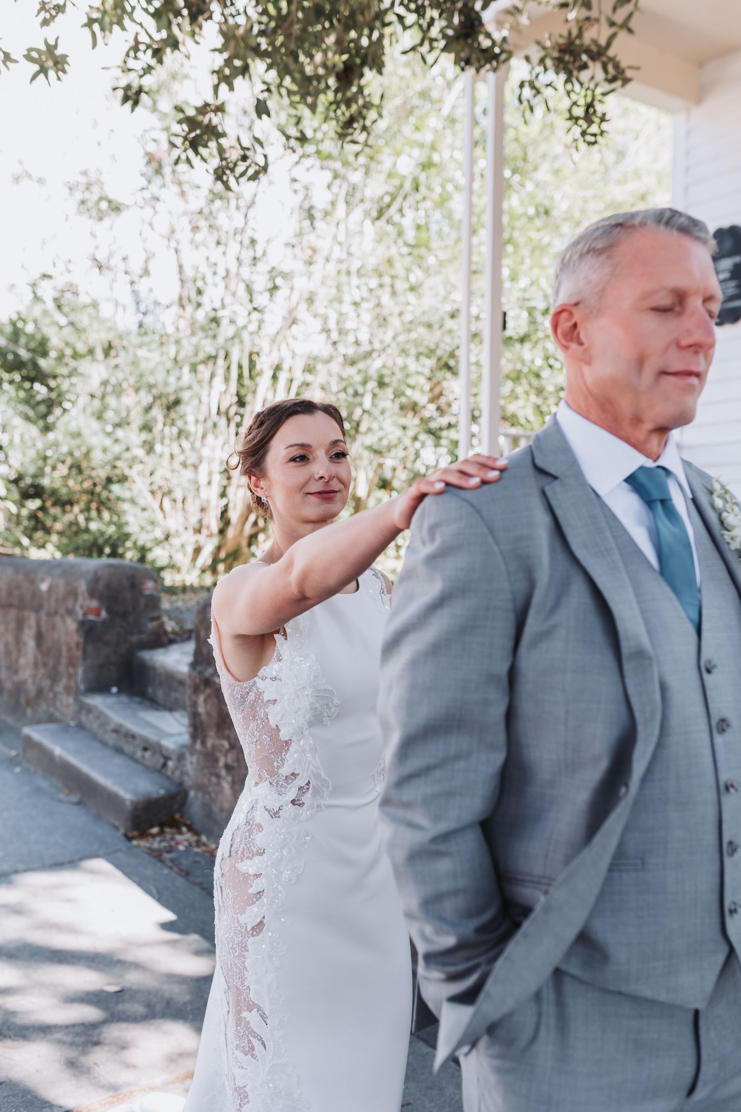 Bride in lace wedding gown tapping groom's shoulder during an emotional first look moment outdoors.