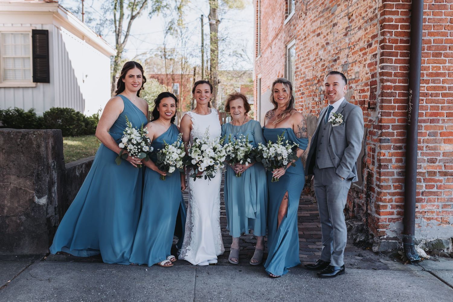 Bride with bridesmaids in dusty blue gowns holding white bouquets beside a brick wall.