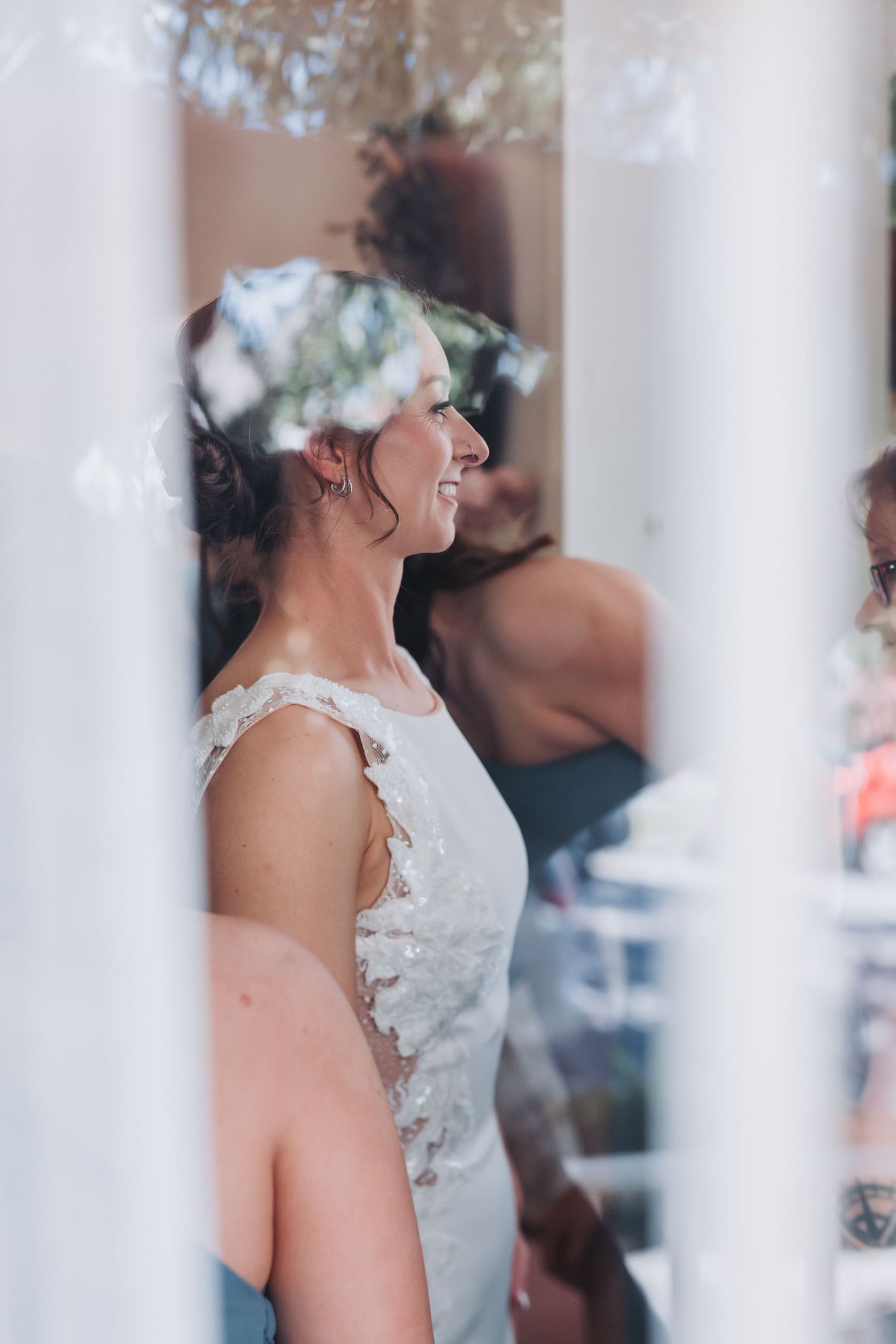 Radiant bride in lace wedding dress smiles joyfully while standing between white columns before ceremony.