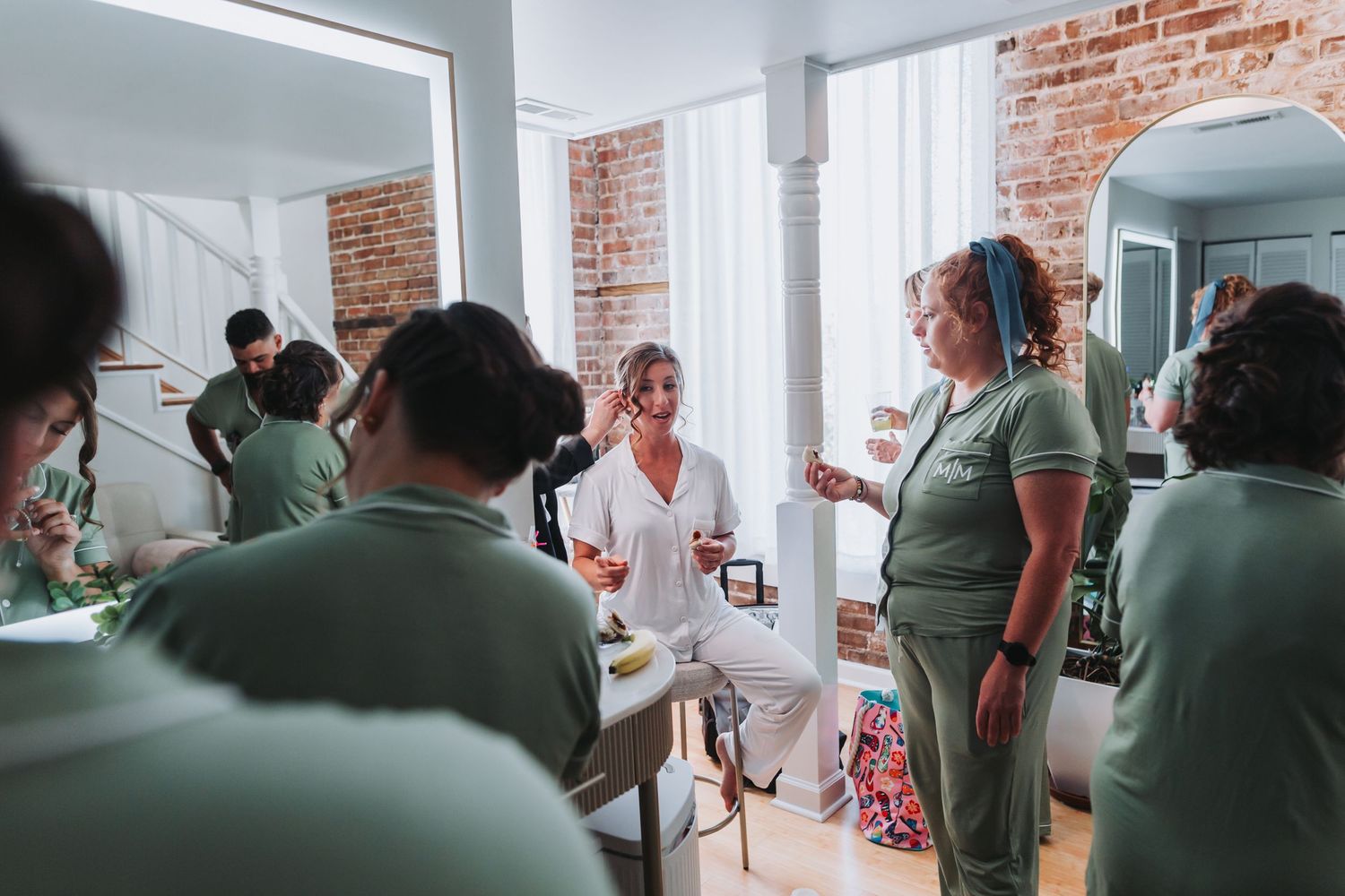Bridal party gets ready in a bright room with exposed brick walls, surrounded by mirrors and natural light.