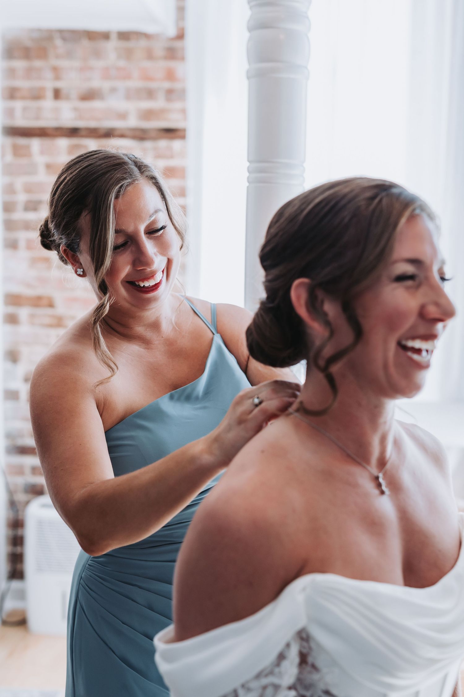 Bridesmaid in sage green dress laughs while helping bride with necklace during wedding preparations indoors.