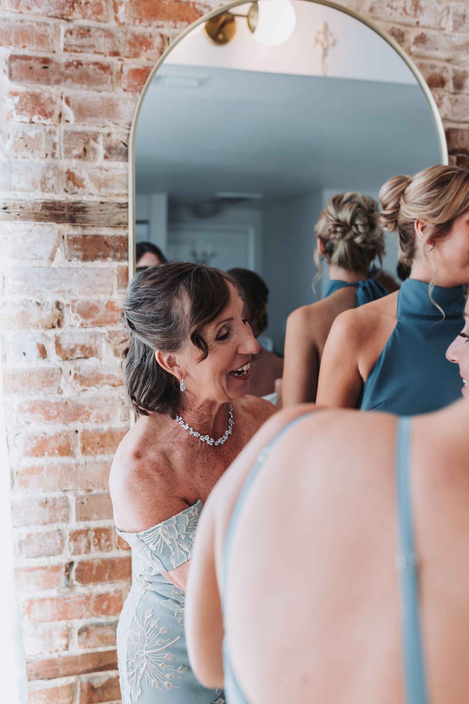 Woman in a blue dress smiling in a mirror reflection during wedding party preparations.