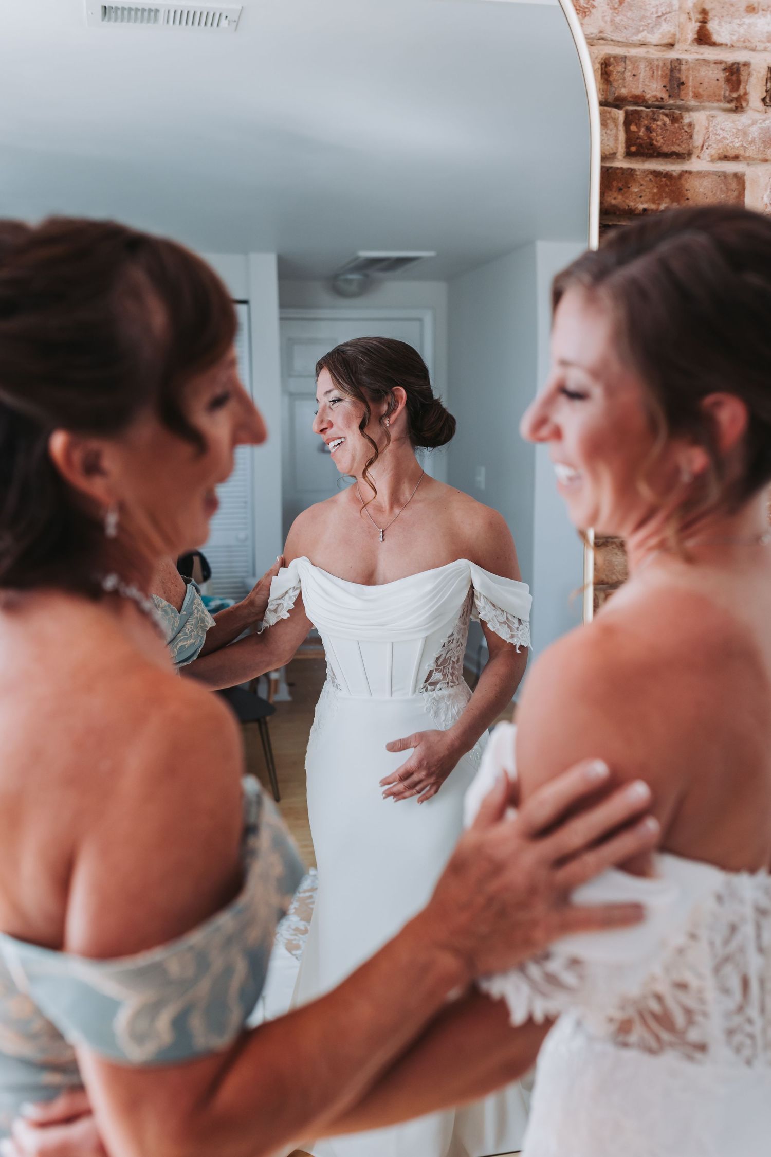 Mother helping bride into off-shoulder white wedding dress reflected in oval mirror.