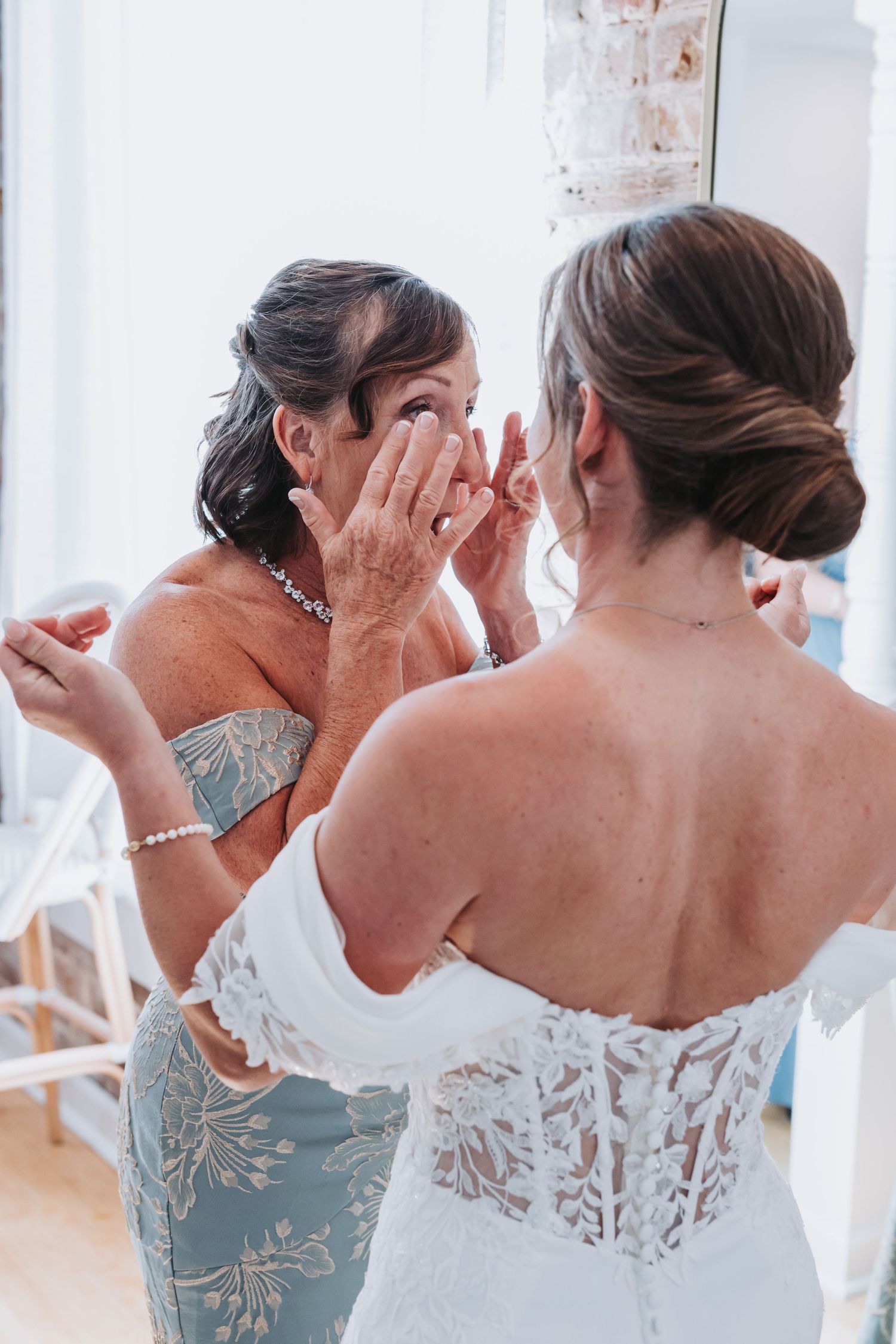 Tearful mother wiping eyes while facing bride in off-shoulder lace wedding gown before ceremony.