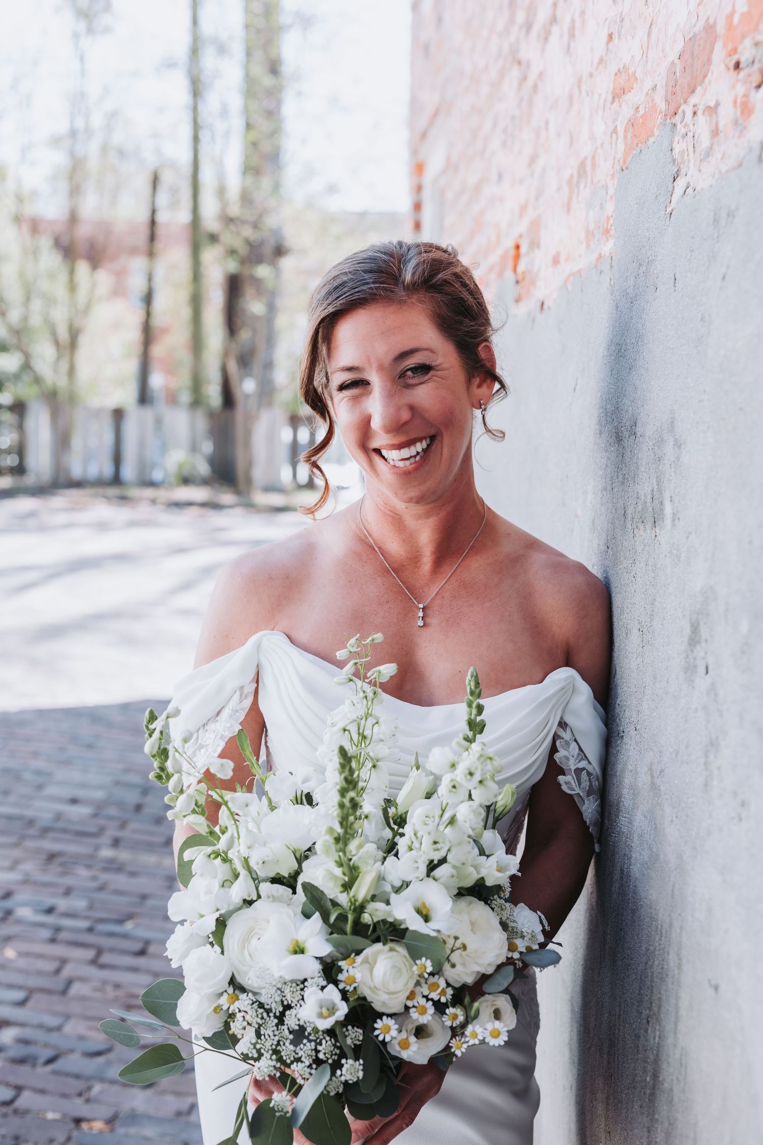 Joyful bride in off-shoulder white gown holding white floral bouquet leaning against brick wall.