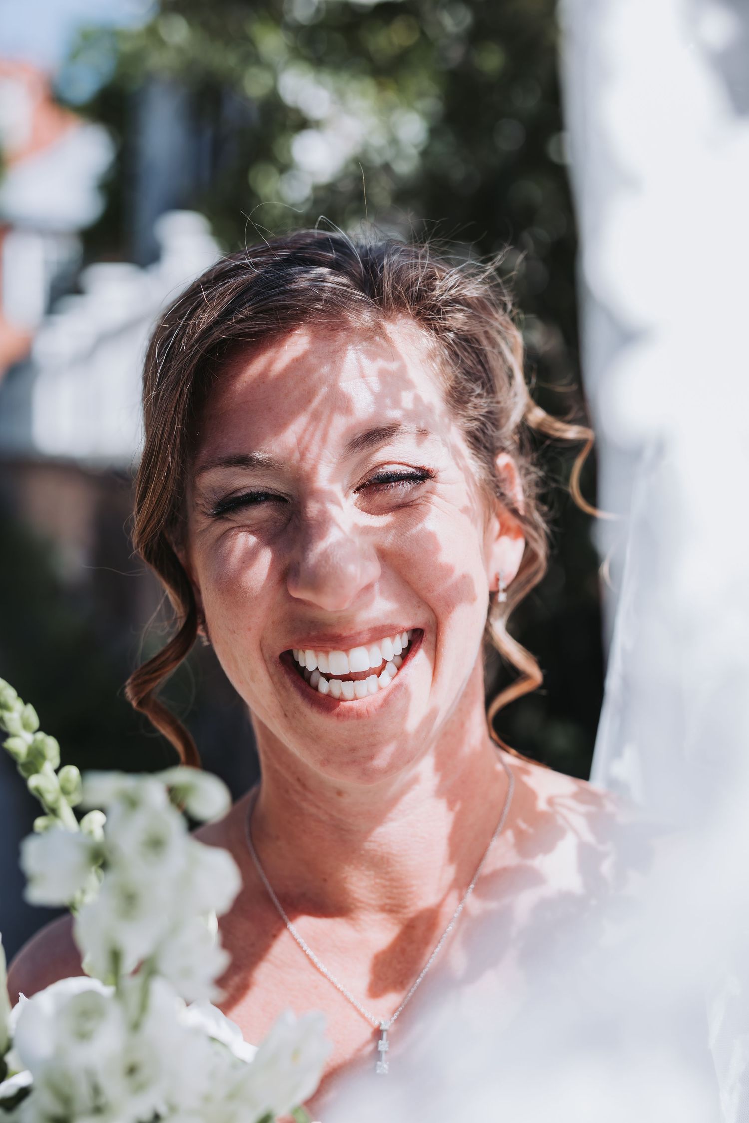 Joyful bride laughing in sunlight with white flowers and veil visible in outdoor wedding portrait.
