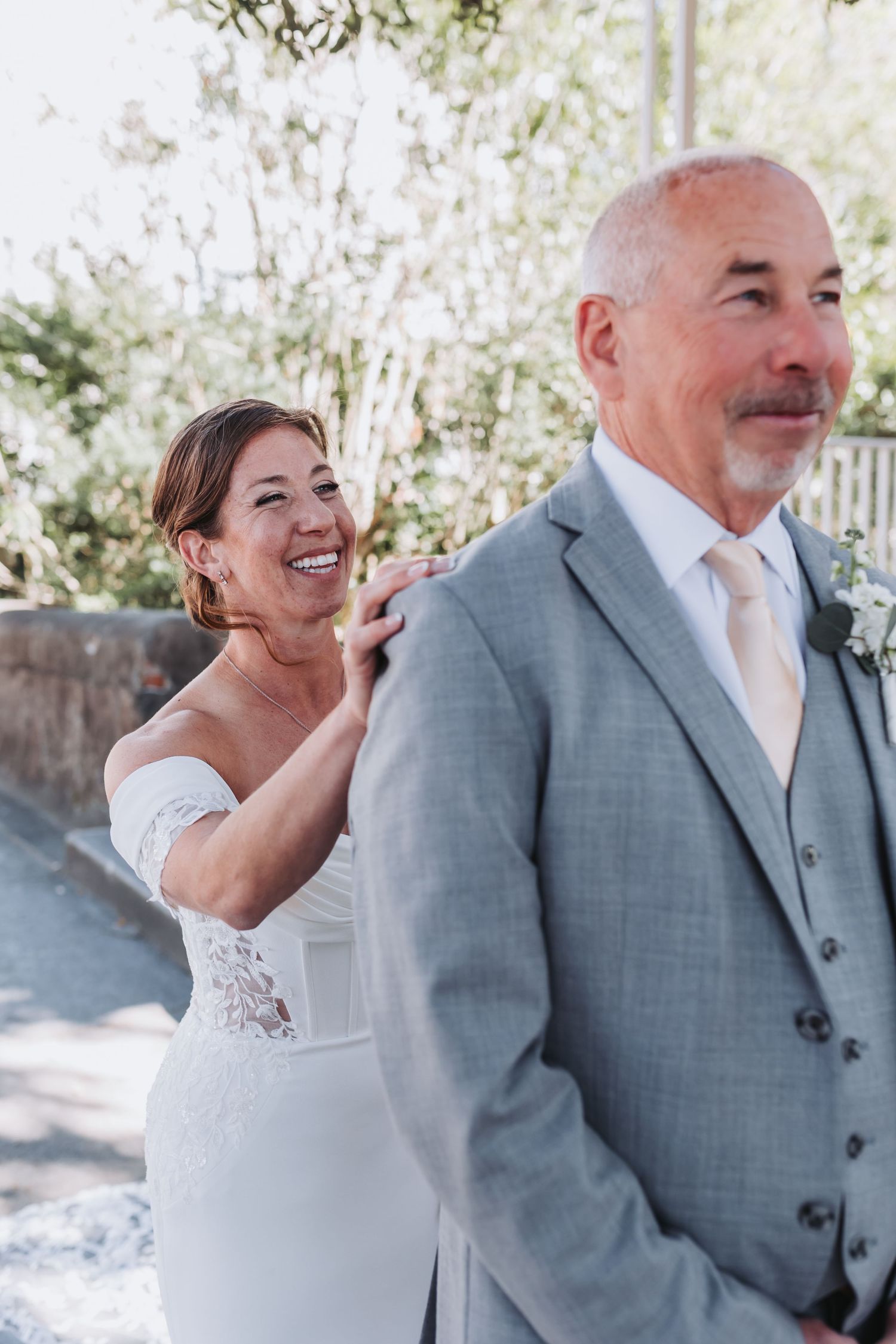 Smiling bride in white lace gown places hand on grooms shoulder during wedding first look moment outdoors.