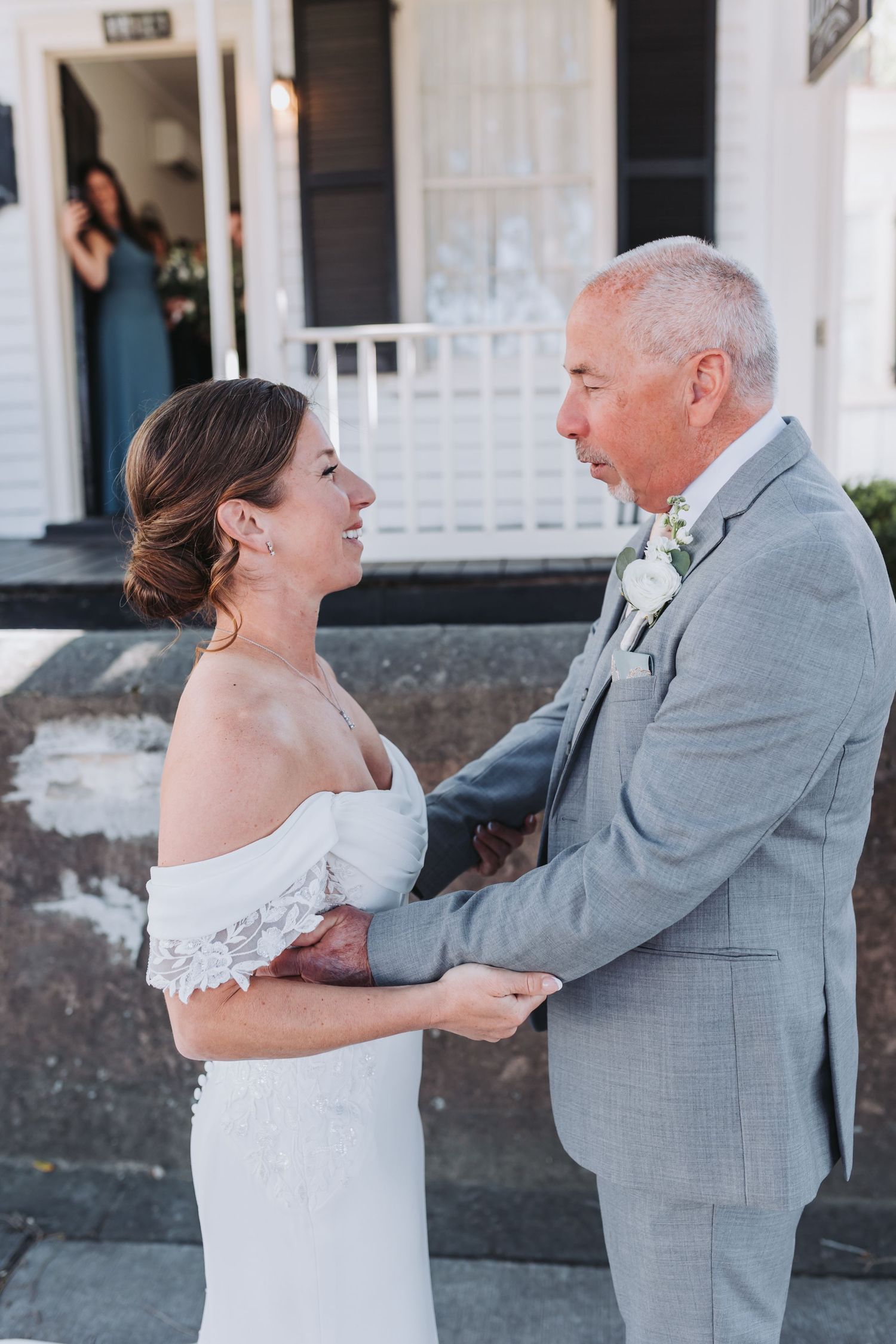 Bride and groom share a tender first look moment, holding hands outside a white building on their wedding day.