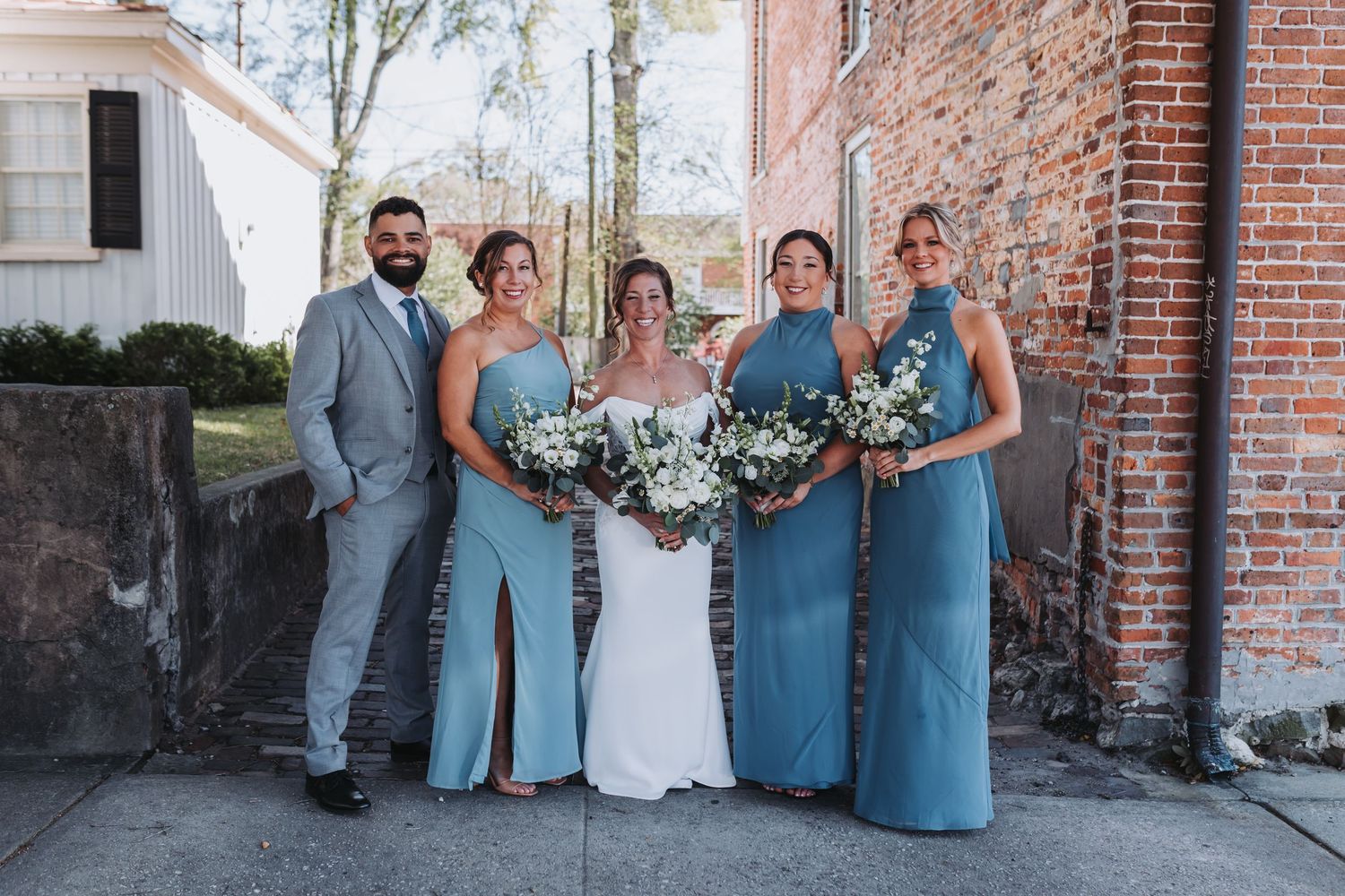 Wedding party posing outdoors with bridesmaids in blue gowns and groomsman in gray suit.