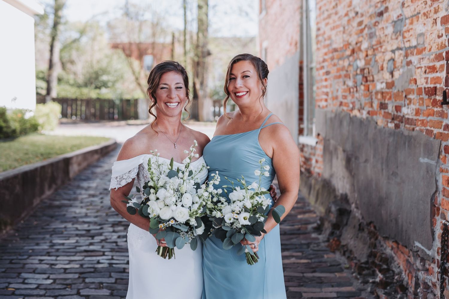 Bride in white off-shoulder gown and bridesmaid in blue dress holding white bouquets outdoors.
