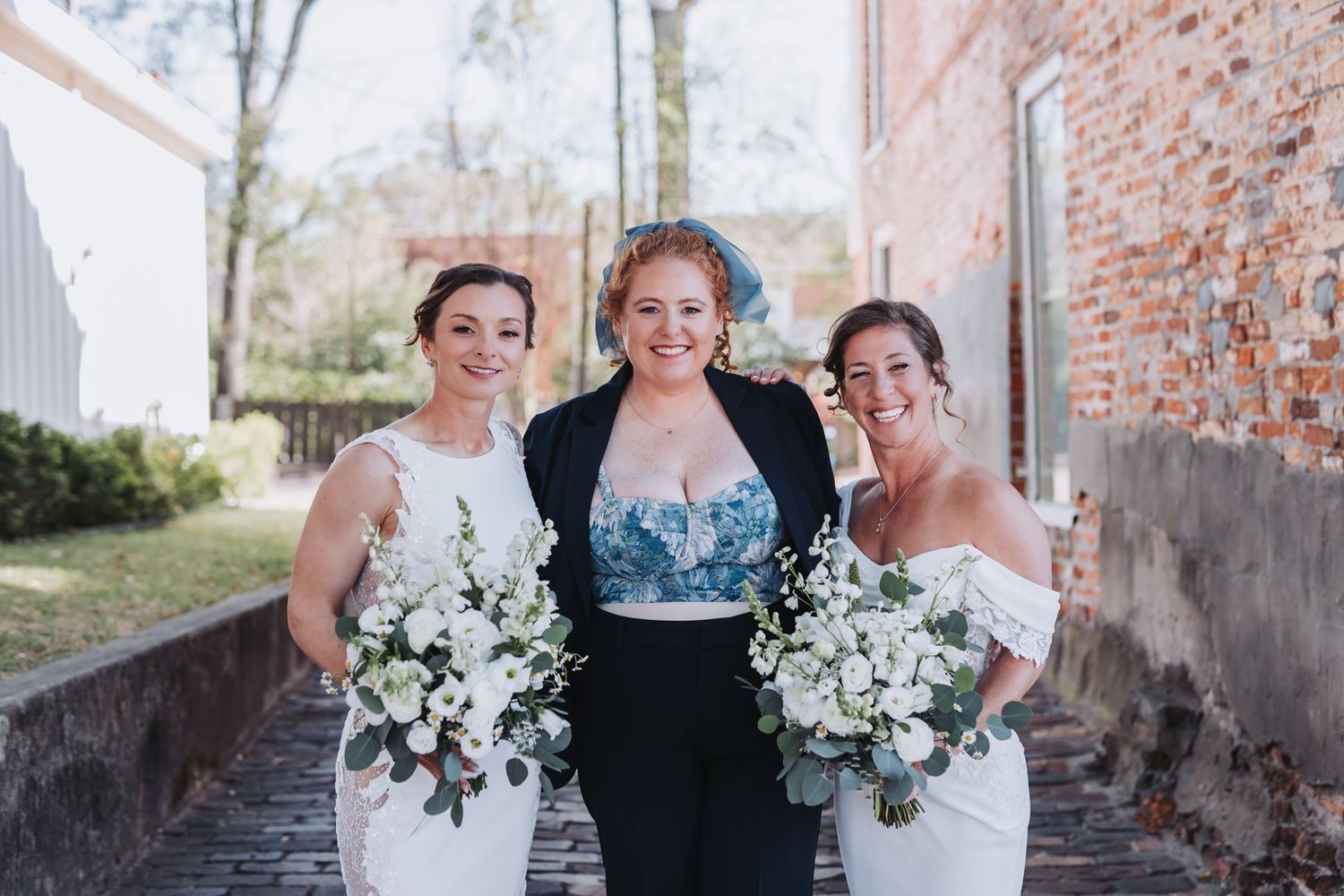 Two brides in white gowns with bouquets flank officiant in blue floral top and black blazer outdoors.