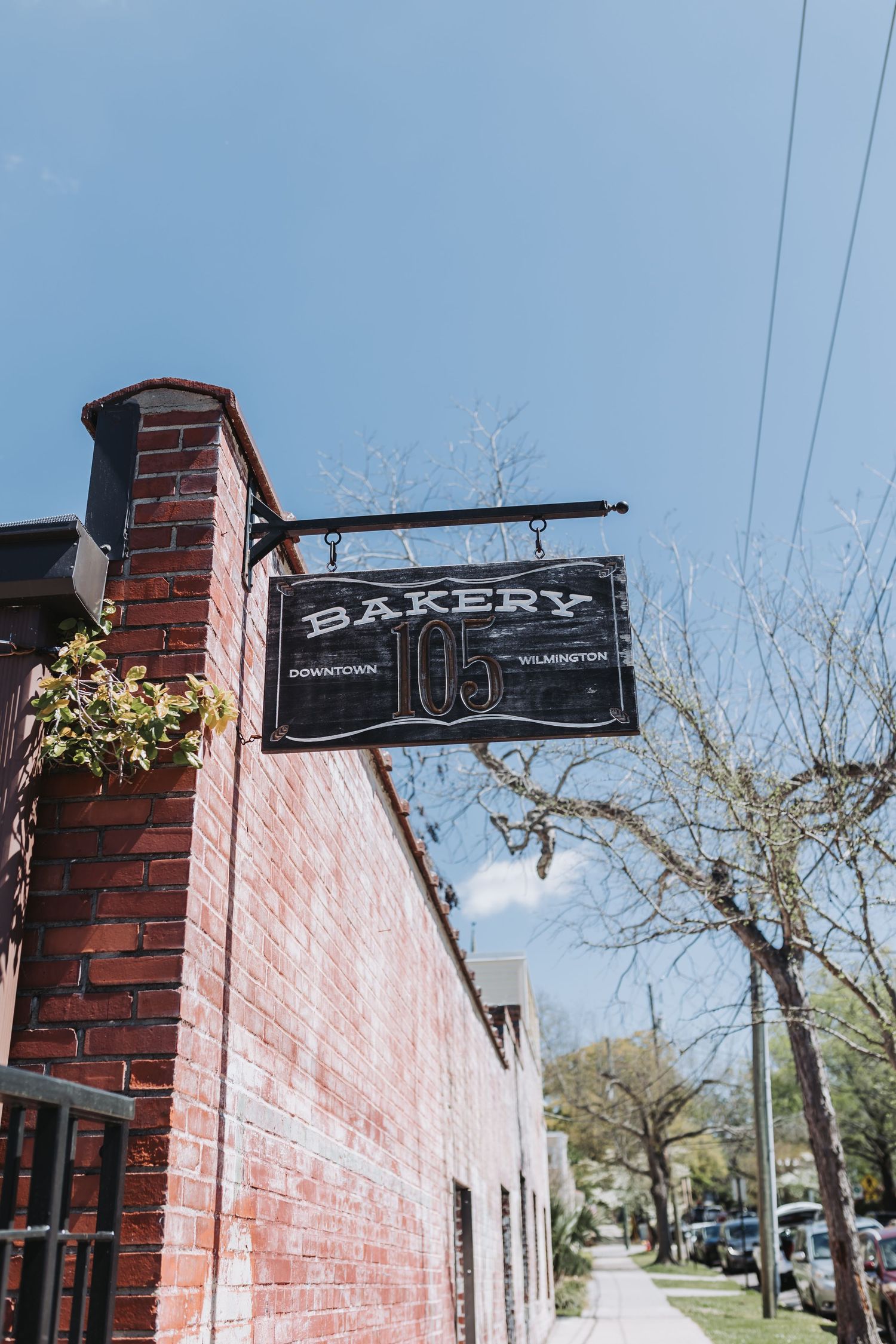 Black bakery sign on brick building in Downtown Wilmington, number 105, against blue sky.
