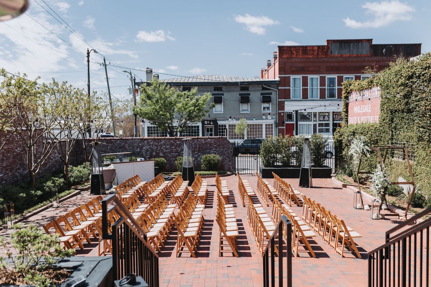 Outdoor wedding ceremony setup with wooden chairs arranged in rows on a brick patio surrounded by lush greenery.
