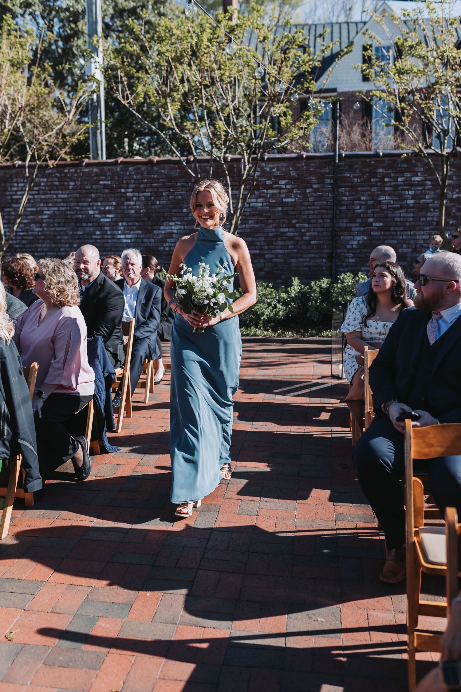 Smiling bridesmaid in teal halter gown walks down brick aisle carrying white bouquet at outdoor wedding ceremony.