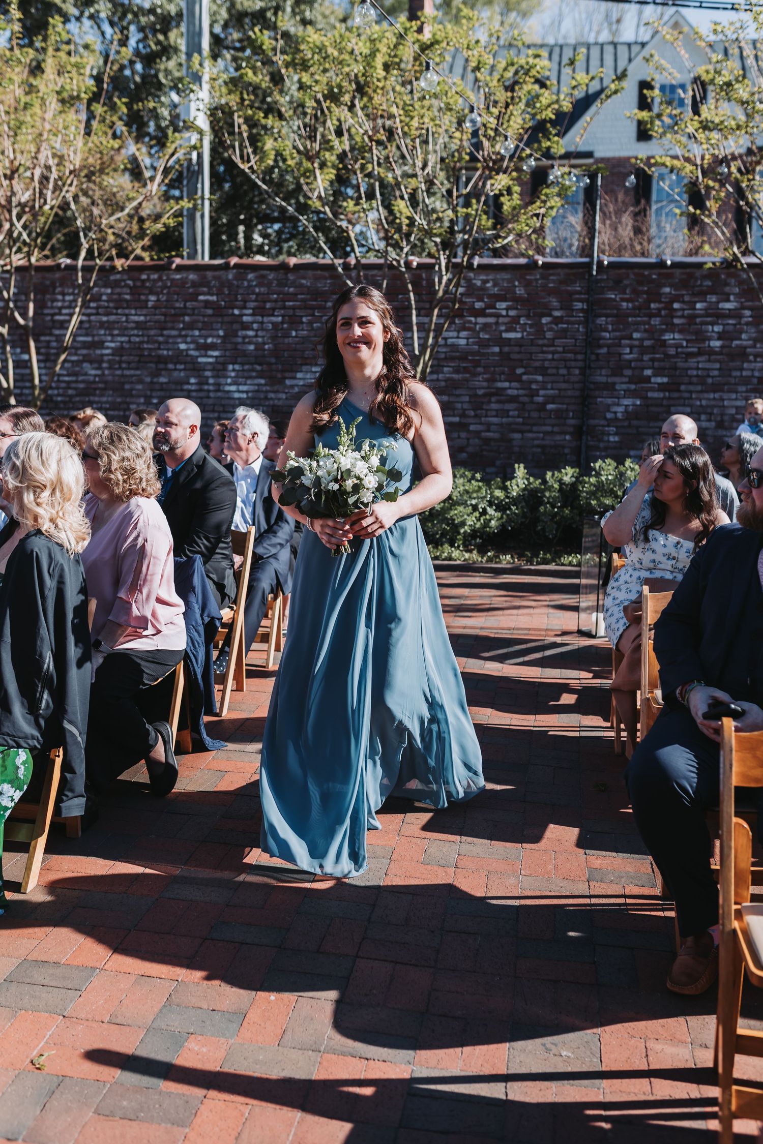 Bridesmaid in blue gown carries white bouquet down brick aisle at sunny outdoor wedding ceremony.