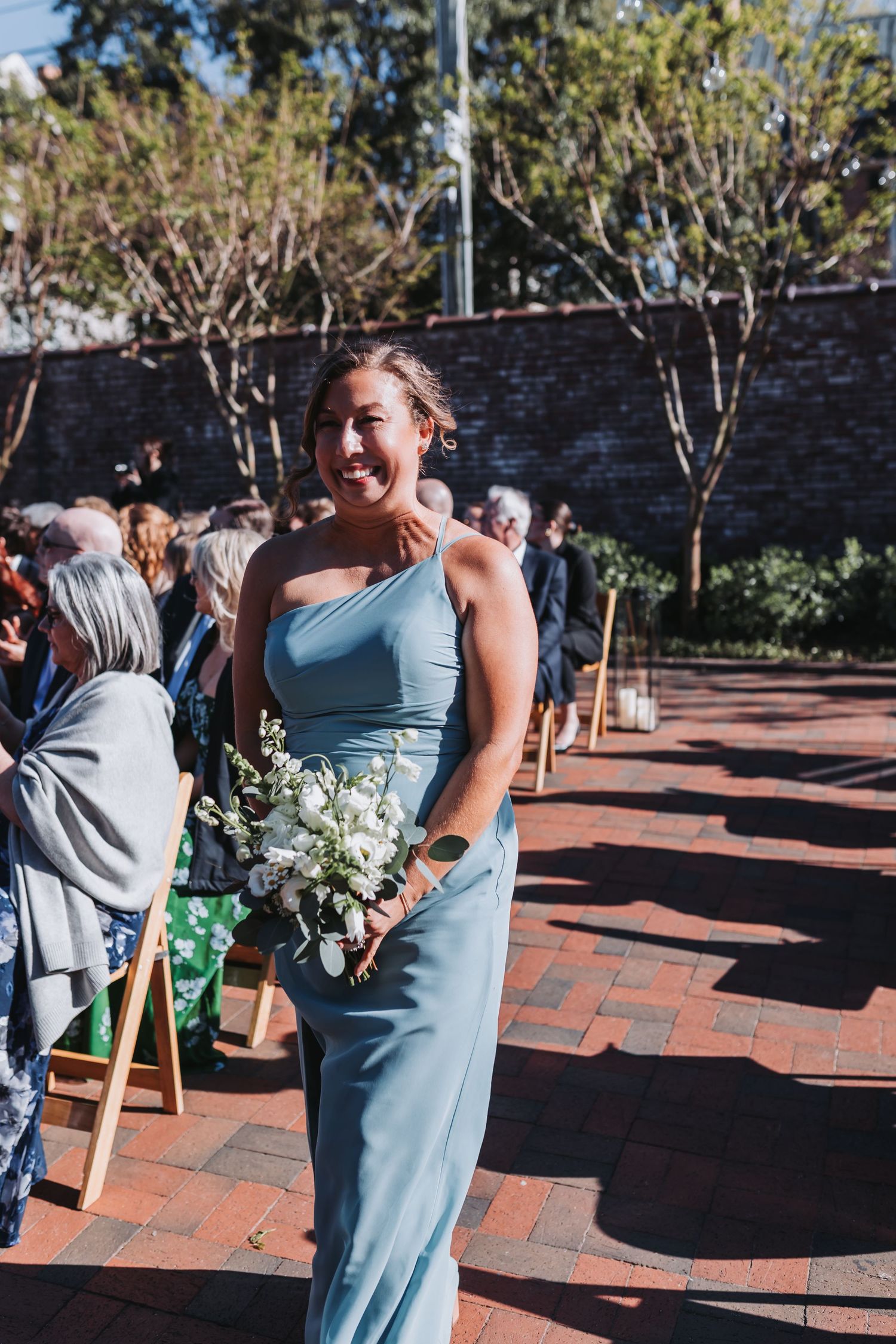 Smiling bridesmaid in blue one-shoulder gown carries white bouquet down brick wedding aisle.