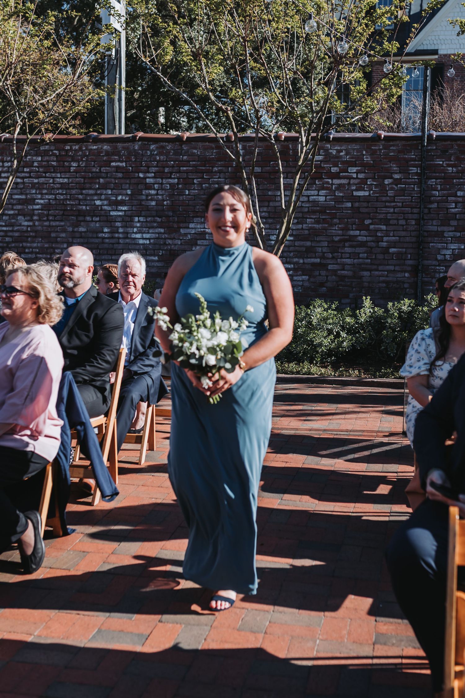Bridesmaid in blue-gray gown walks down brick aisle holding white bouquet at outdoor wedding ceremony.
