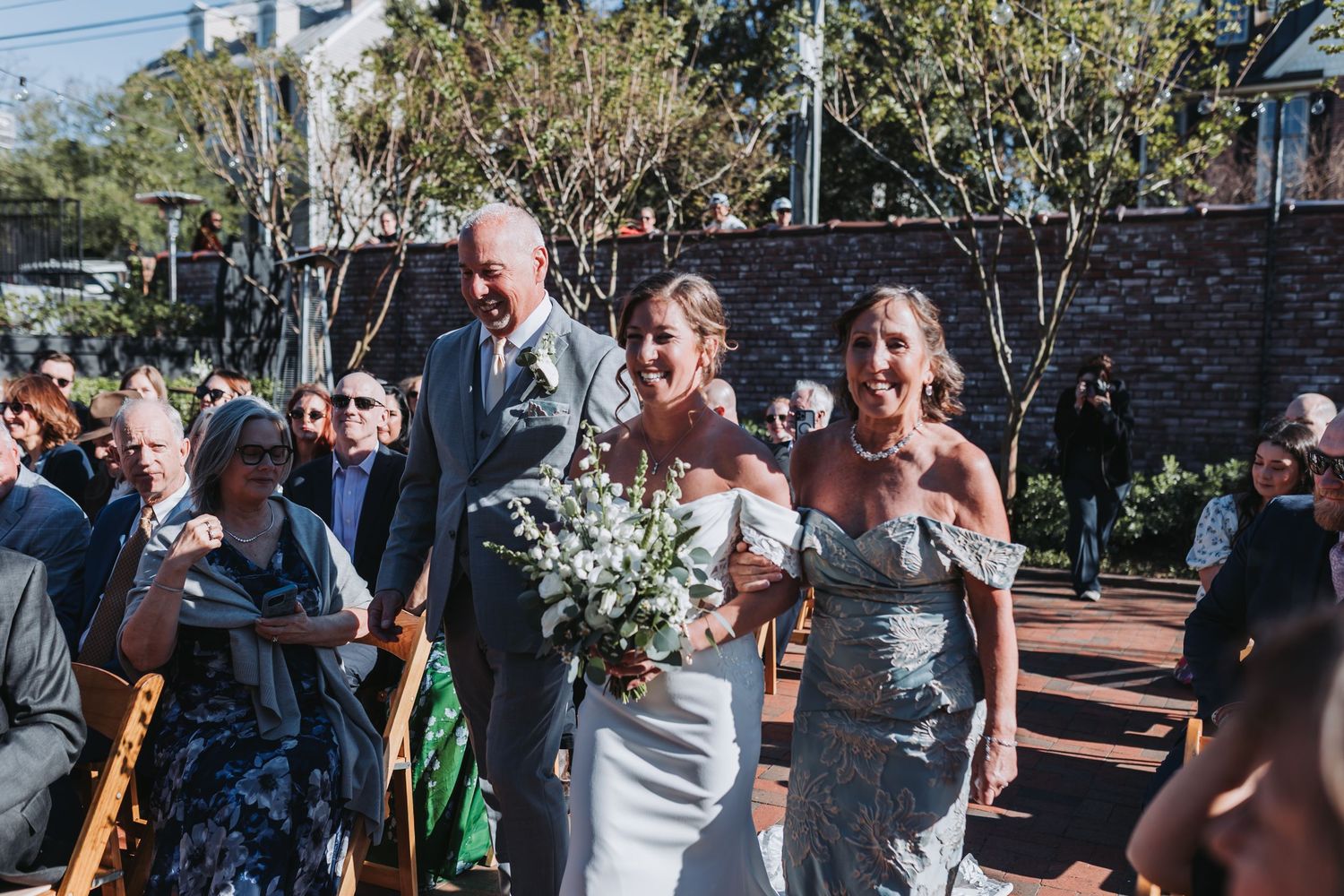 Bride walks down outdoor wedding aisle with two escorts, holding white bouquet, guests seated on either side.