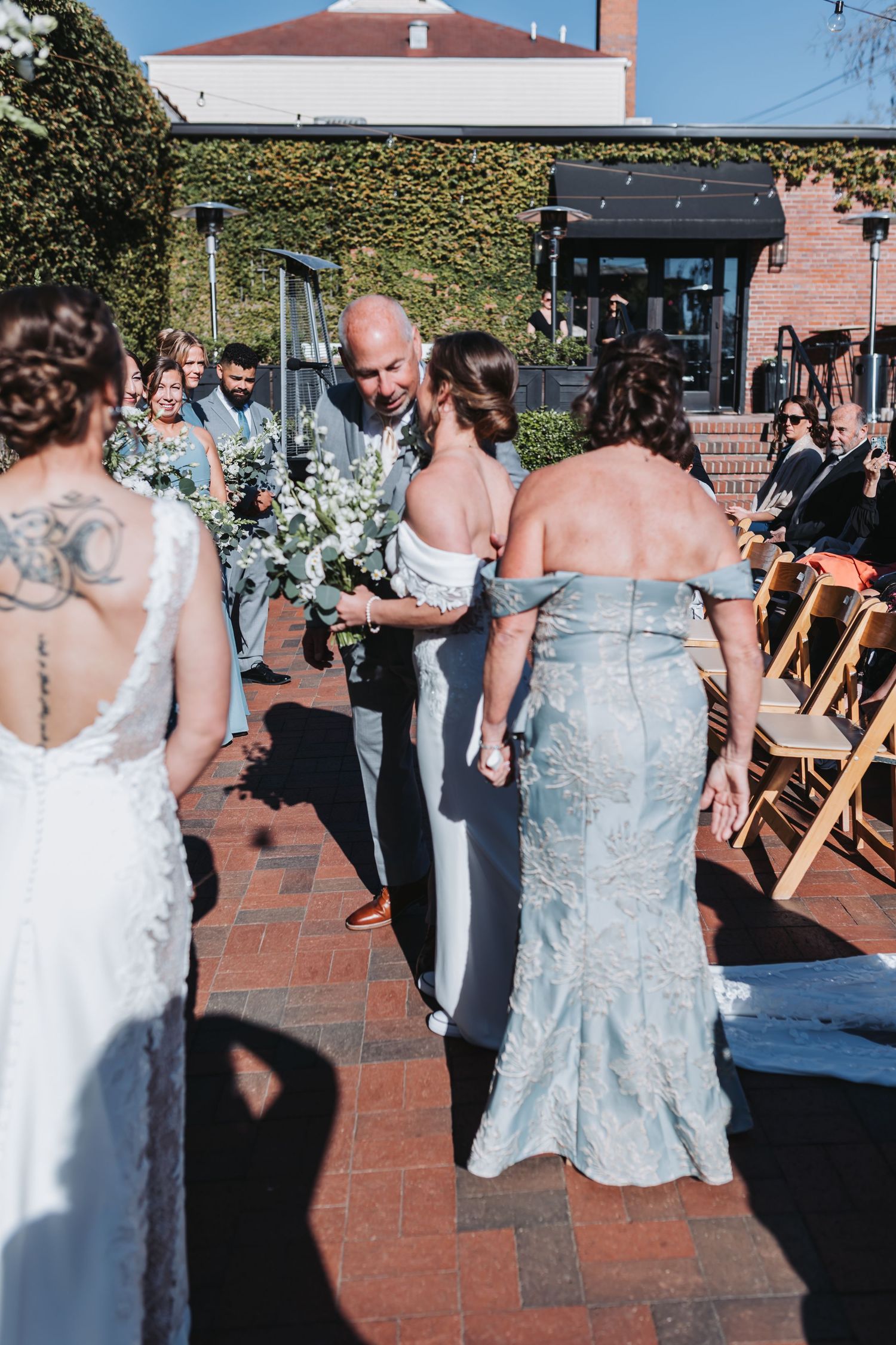 Bald groom in gray suit greets bride and mother at outdoor wedding ceremony on brick patio with ivy wall backdrop.