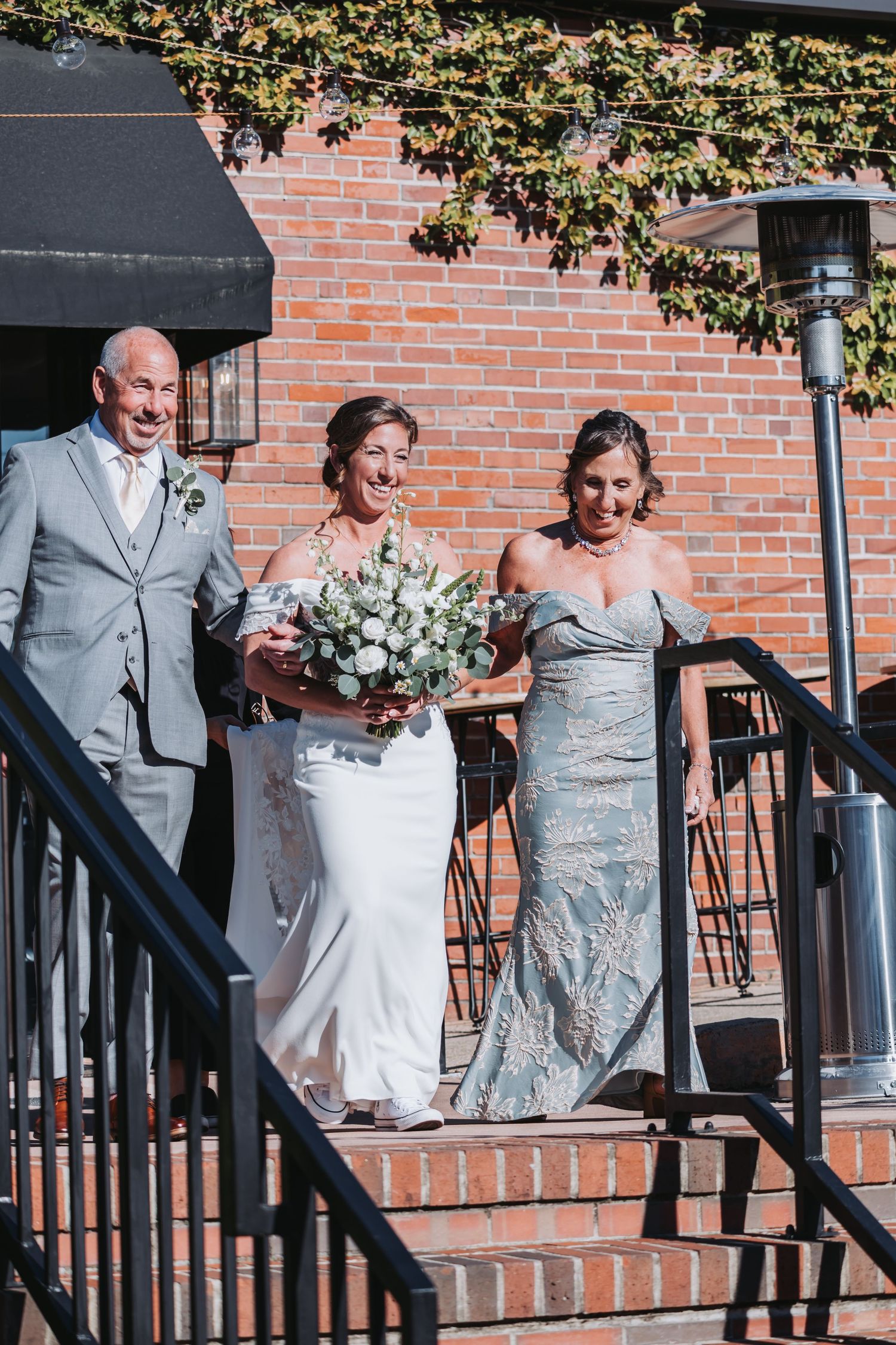 Smiling bride walks down steps with father and bridesmaid holding white bouquet at outdoor wedding.